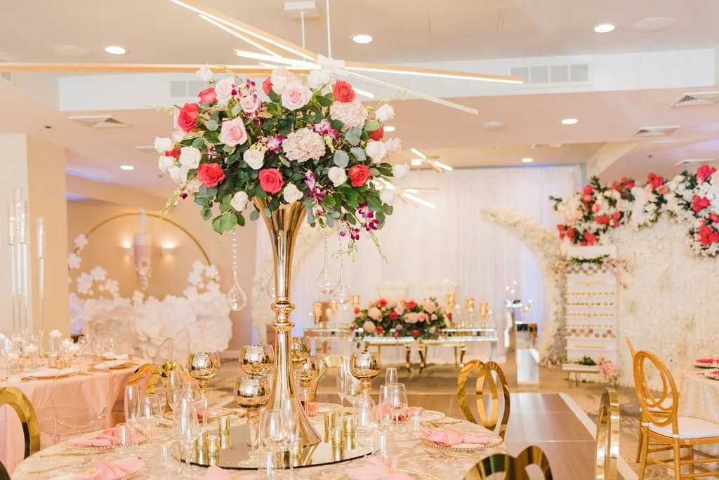 Elegant banquet hall decorated for a celebration with a large floral centerpiece featuring pink, red, and white roses on a tall gold stand, surrounded by glassware, gold accents, and pink napkins.