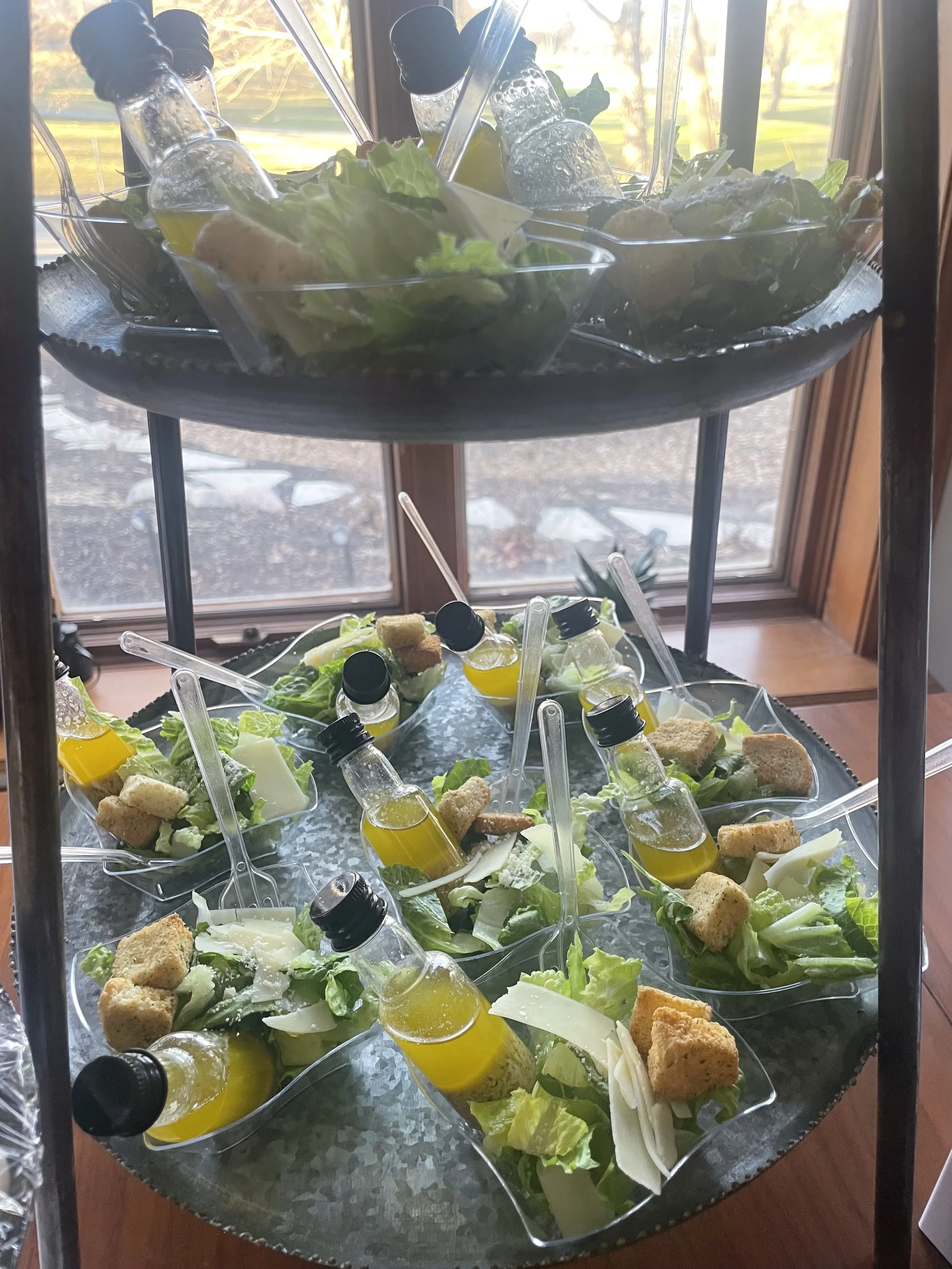 Tray of individual salads with lettuce, cheese, croutons, and small bottles of dressing, arranged on a two-tiered serving stand near a window.