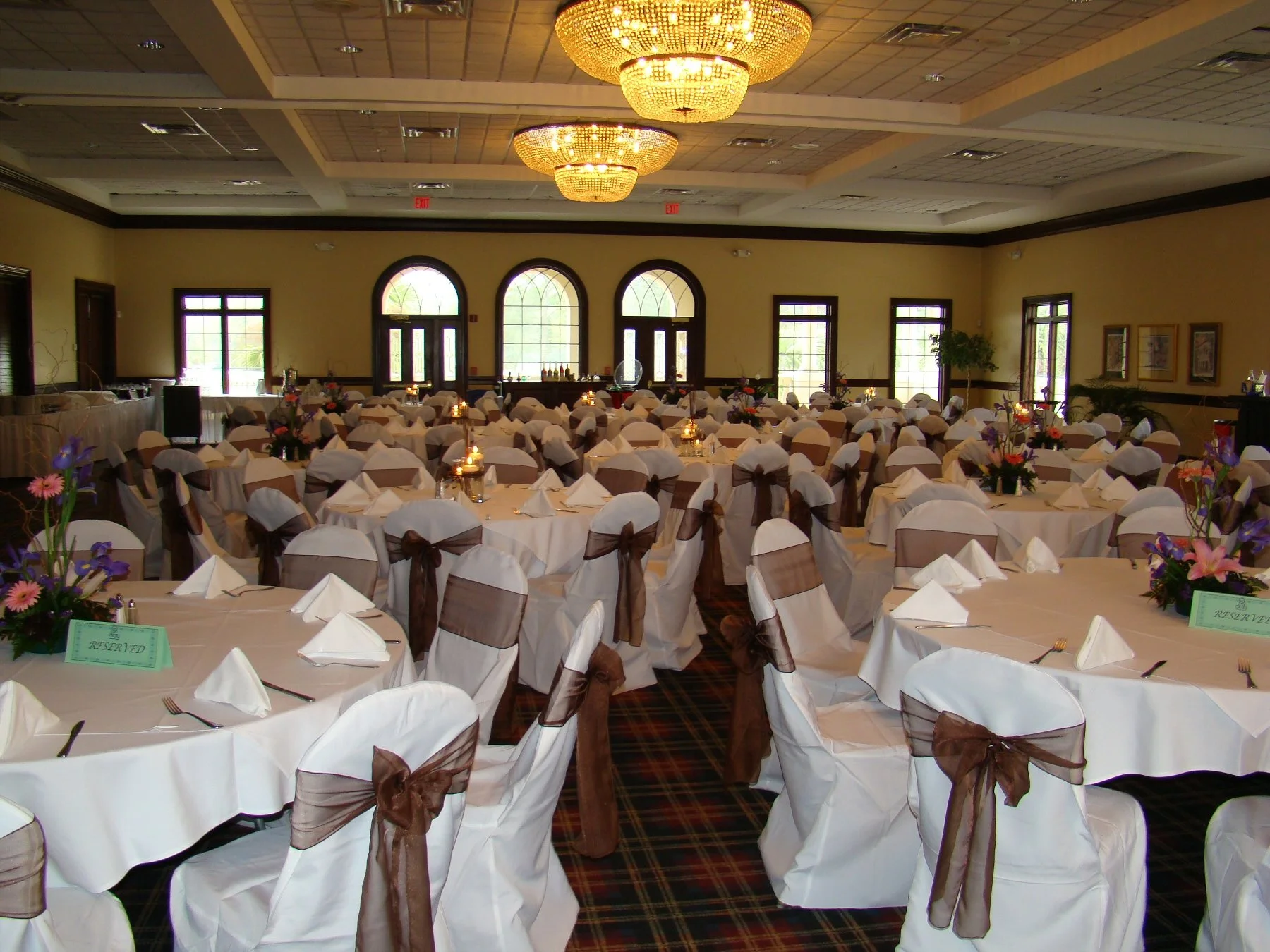 A banquet hall decorated for an event with round tables covered with white tablecloths, each set with folded napkins and cutlery. Chairs are draped in white fabric with brown ribbons tied in bows. There are floral centerpieces on the tables and reser