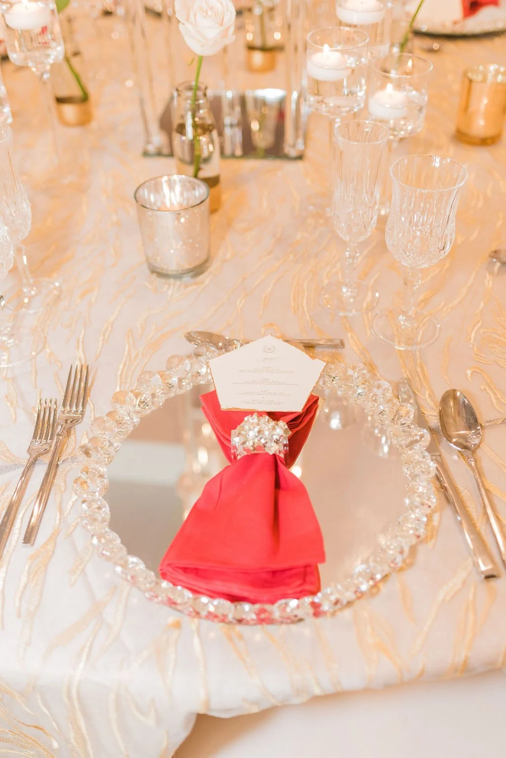 Elegant wedding table setting with a crystal-edged plate, red napkin, menu card, silverware, and decorative glassware, with a flower in the background.