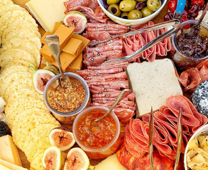 Close-up of a charcuterie board with assorted cheeses, sliced cured meats, figs, olives, jam, mustard, and crackers.