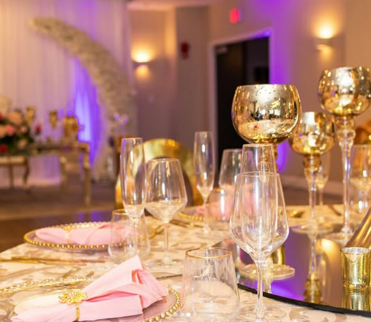 Elegant event table setup with pink napkins, gold accents, and reflective gold candle holders with lit candles, in a decorated venue with purple and warm lighting.