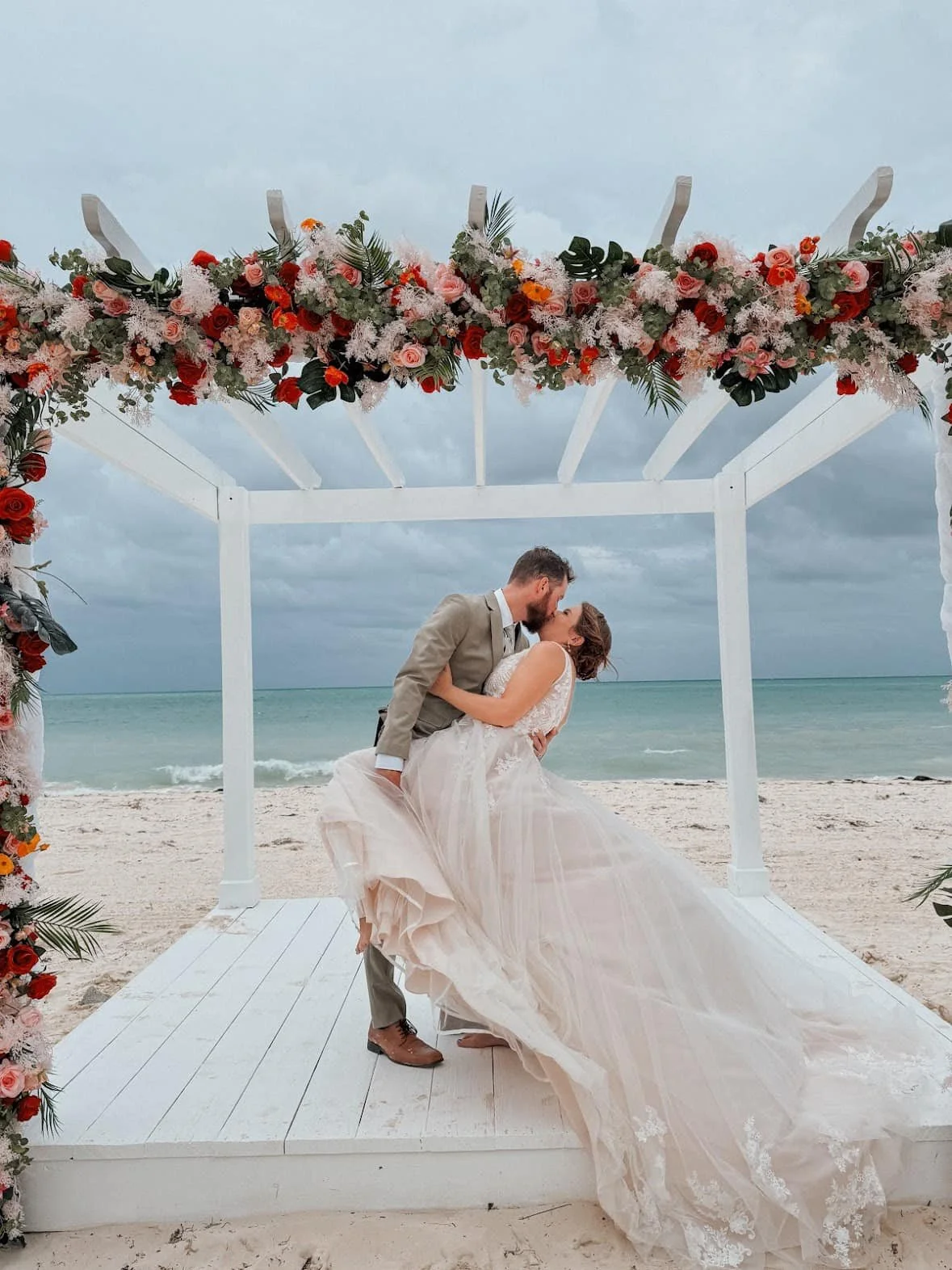 A couple in wedding attire sharing a kiss on a beach under a floral arch with gray sky and ocean in the background.