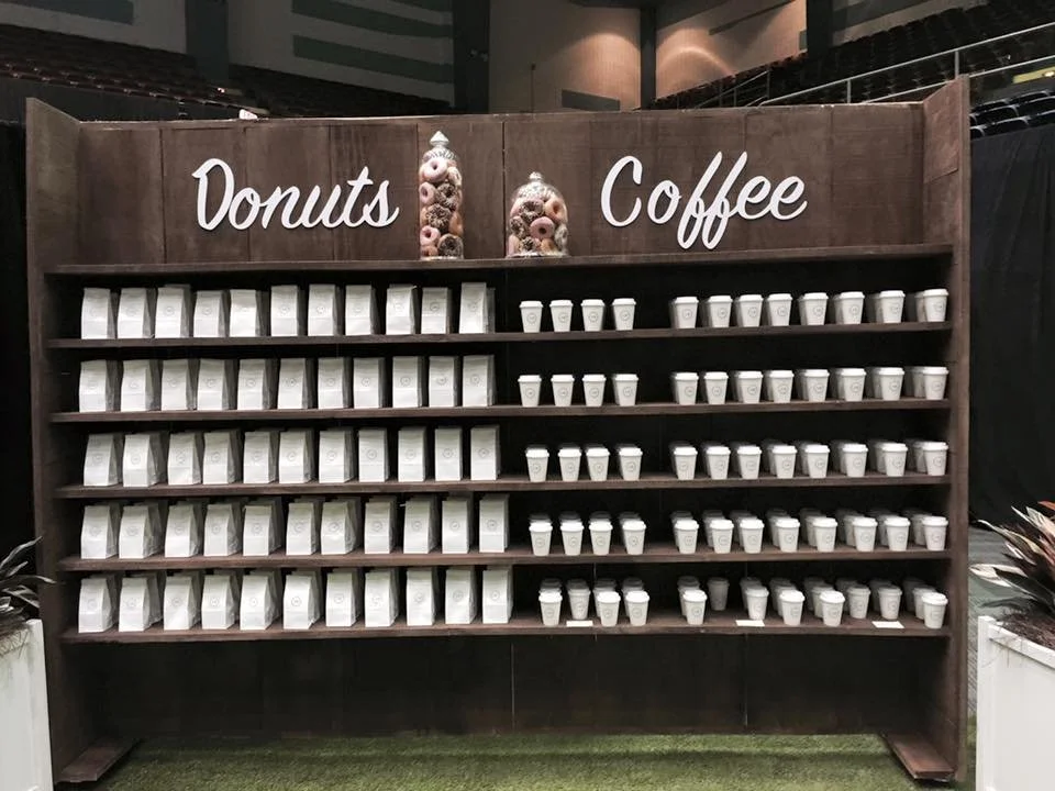 Display shelf with multiple white bags of donuts on the left and white coffee cups on the right, with 'Donuts' and 'Coffee' written at the top, and decorative jars with donuts.