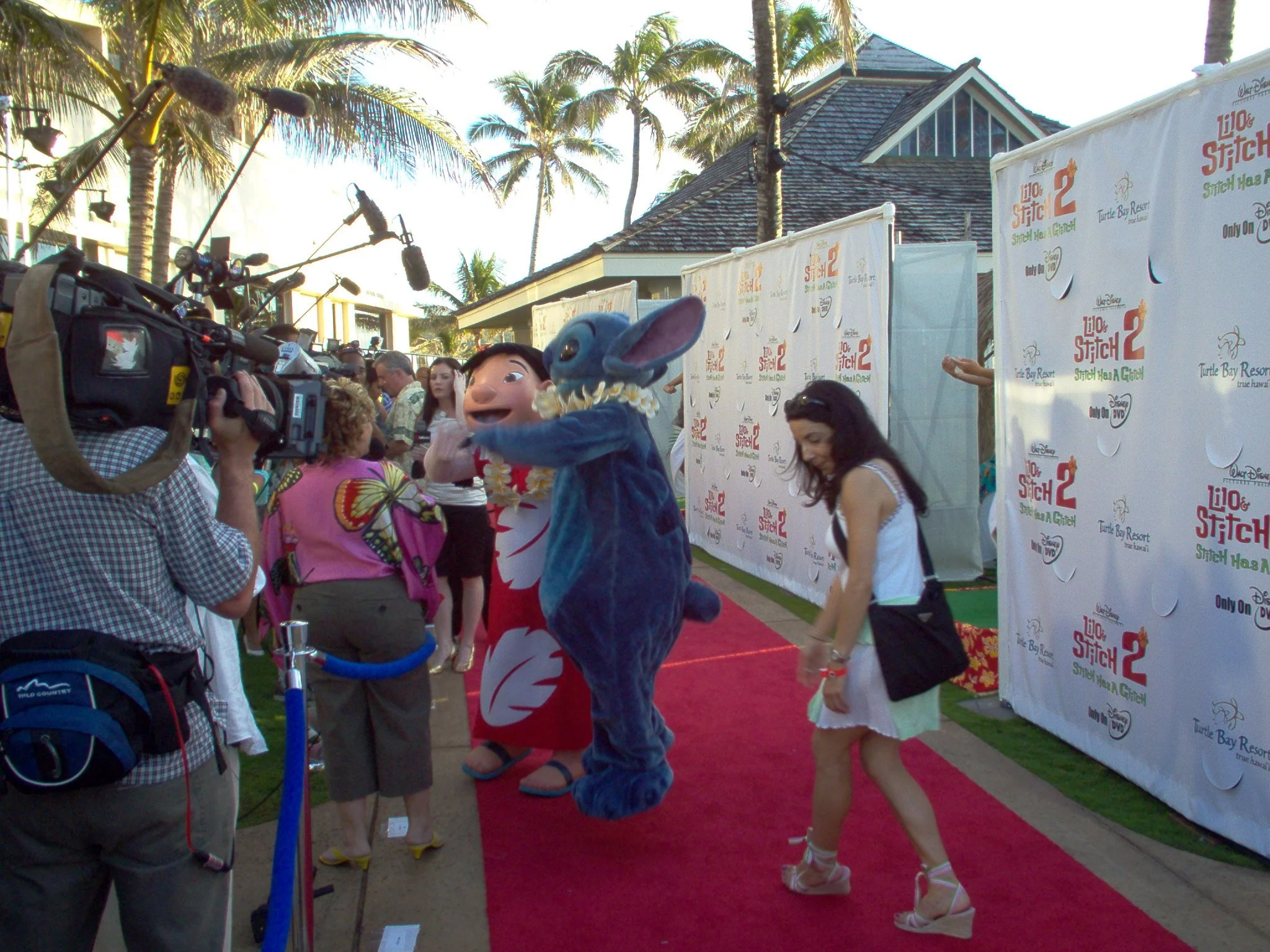 Characters and mascots on a red carpet at a Disney event, with a person dressed as Stitch from Lilo & Stitch, and a woman in a white dress, with media and onlookers observing.
