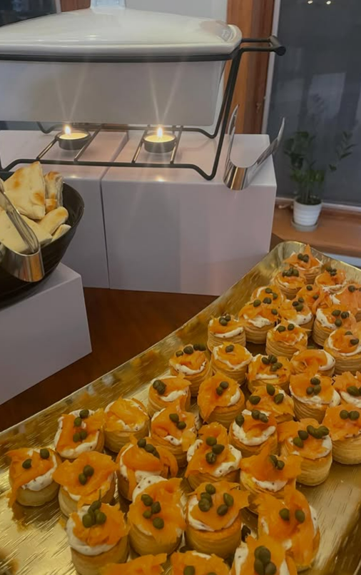 Tray of small appetizers topped with cream, smoked salmon, and green peppercorns, arranged on a golden platter at a buffet table.