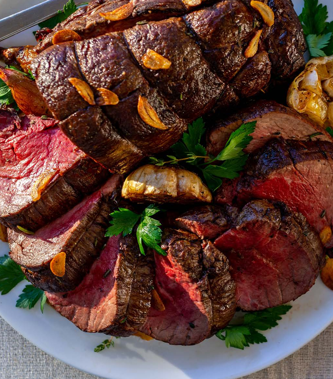 Sliced medium-rare roast beef garnished with parsley and garlic cloves on a white plate.