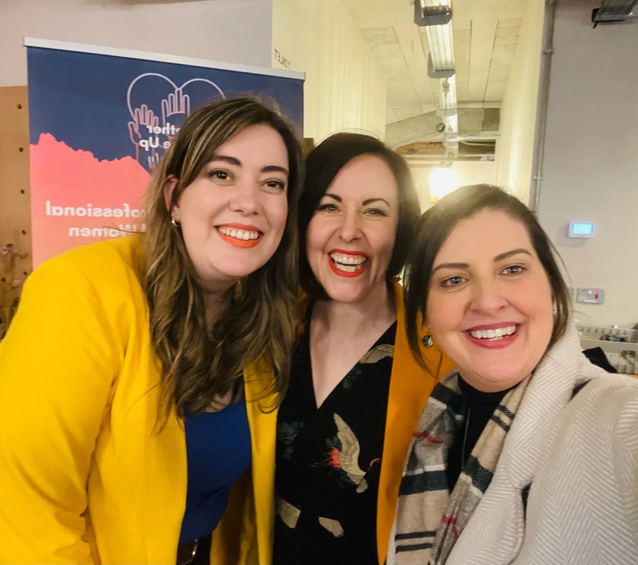 Three women smiling and taking a selfie at an indoor event with a colorful banner in the background.
