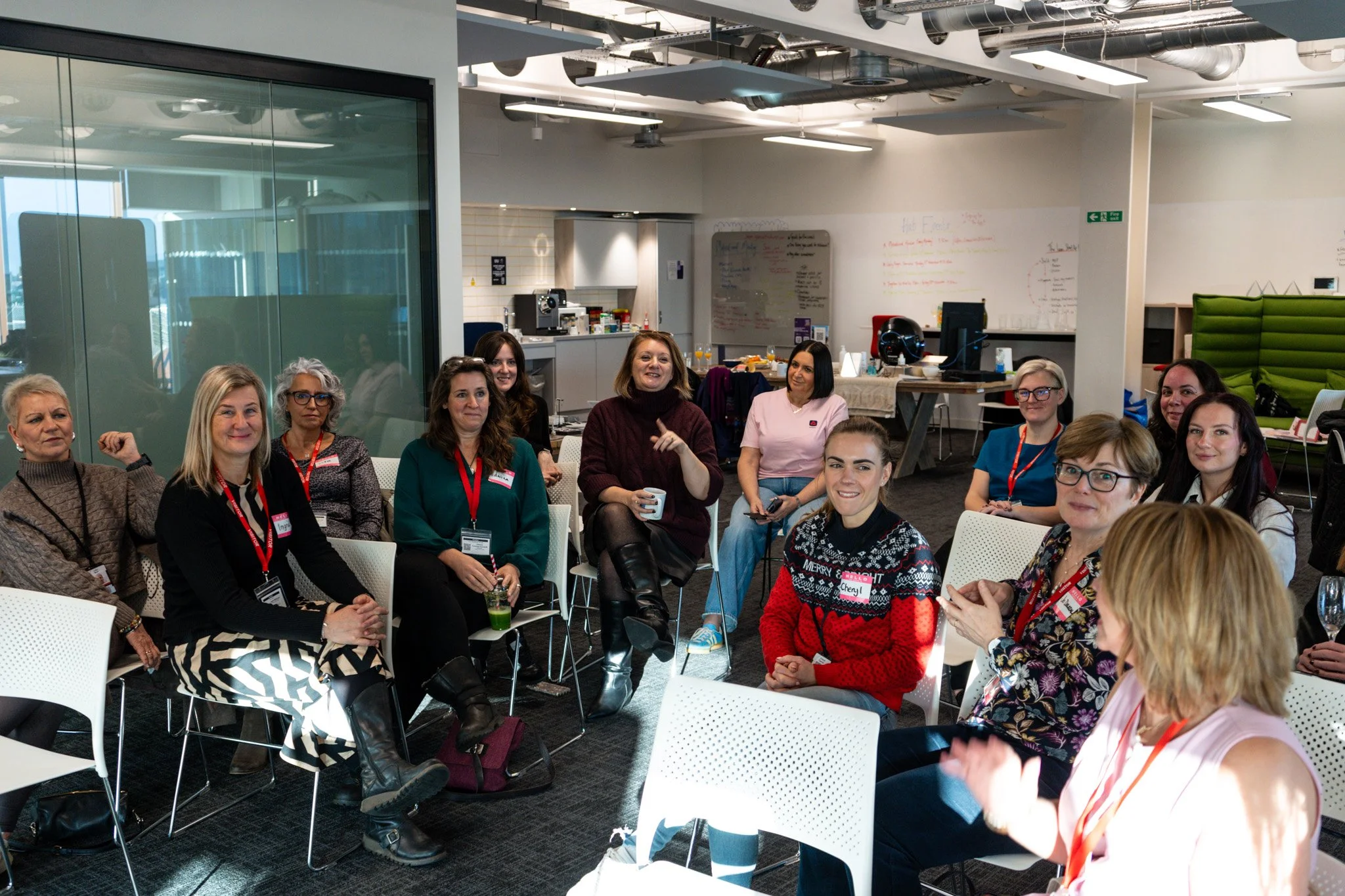 Group of women sitting in a conference room, engaging in a discussion, with some holding drinks, and whiteboards and kitchen area in the background.