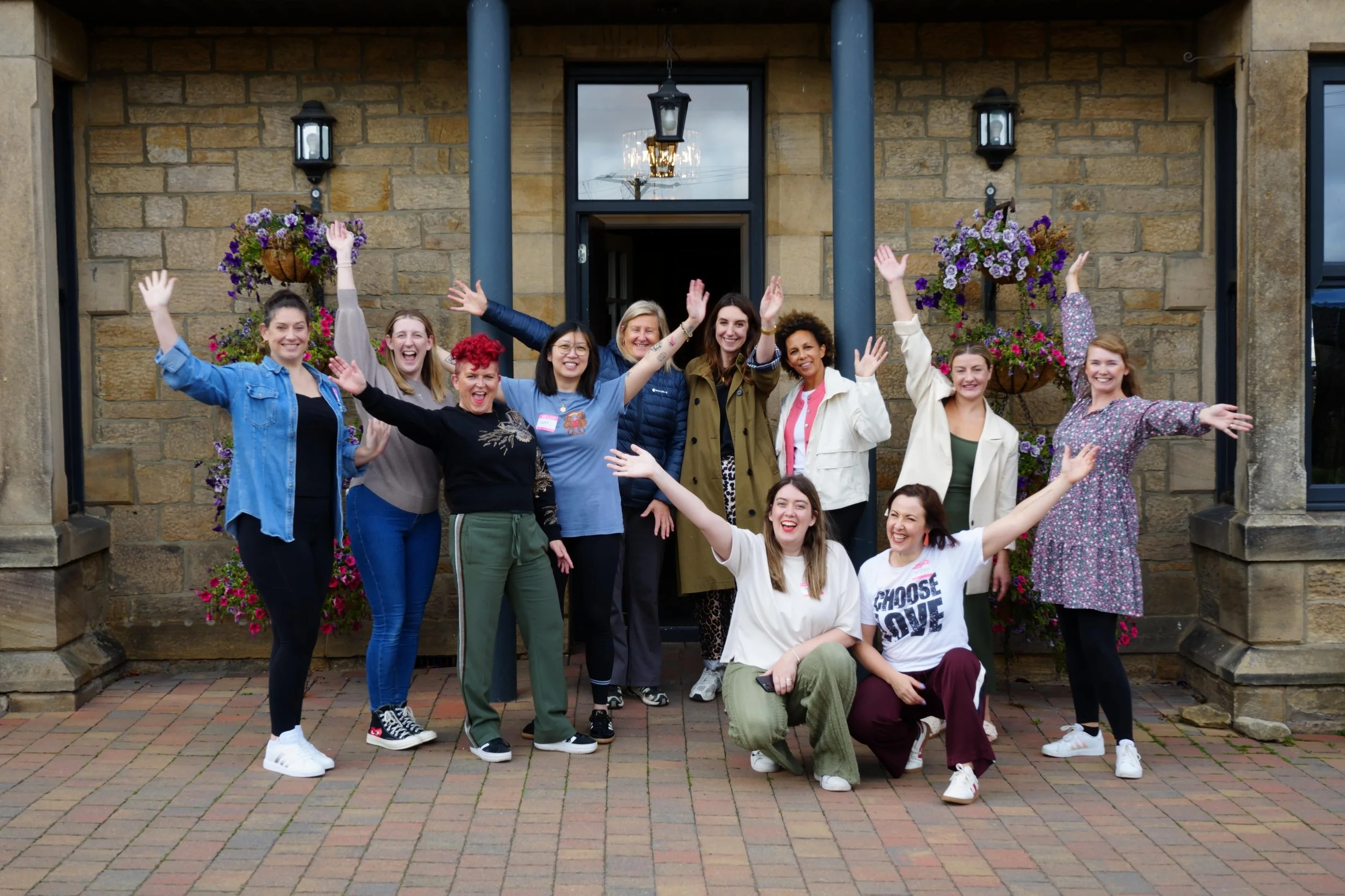 A group of women posing cheerfully in front of a brick building with hanging flower baskets, some with arms raised, smiling.