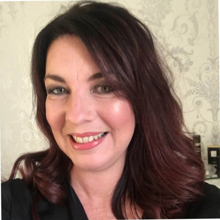 A woman with shoulder-length dark red hair smiling and sticking her tongue out, wearing a black top against a light-colored patterned background.