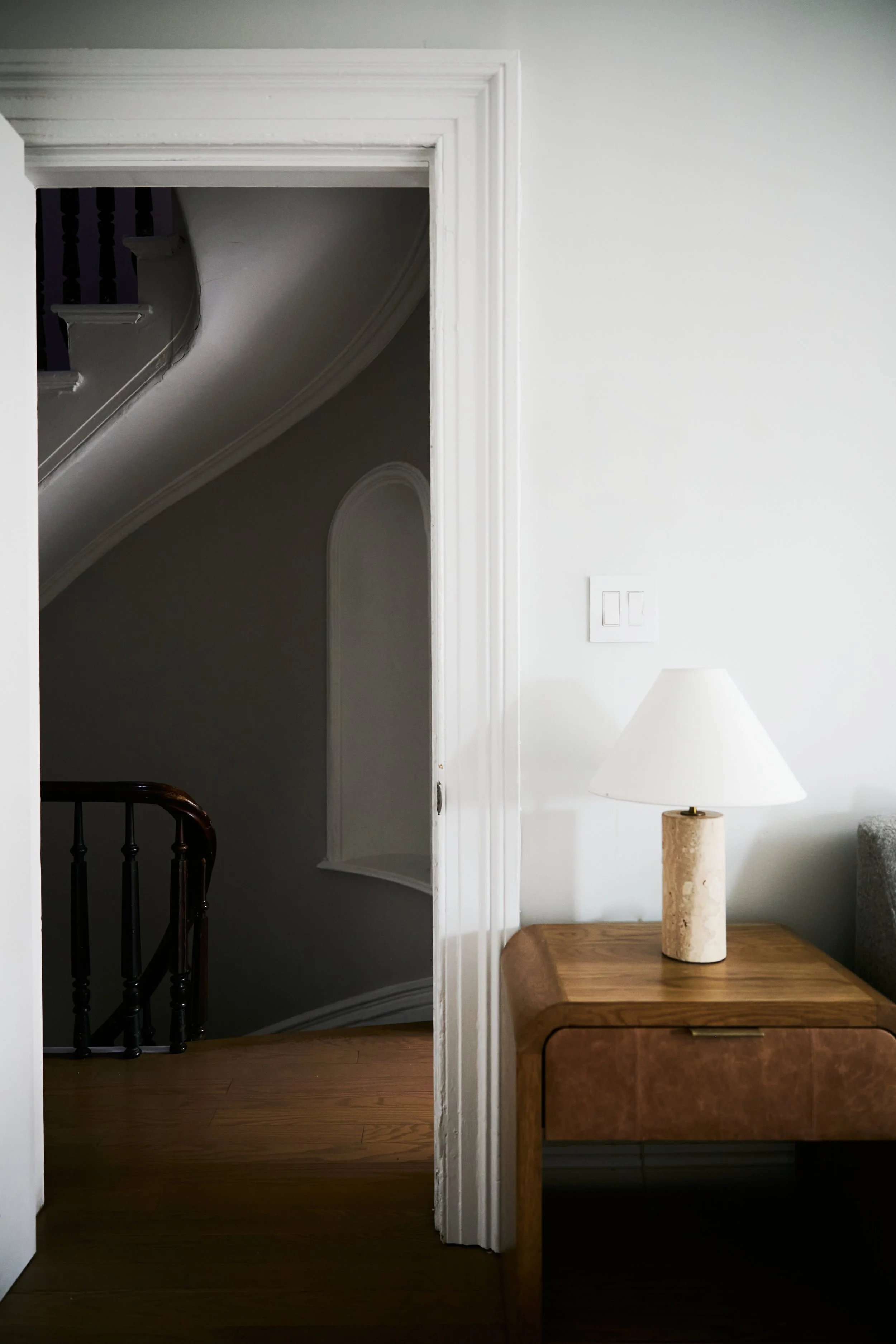 Interior view of a home featuring a wooden table with a beige lamp on it, a white door frame, and a staircase with dark-painted spindles visible in the background.