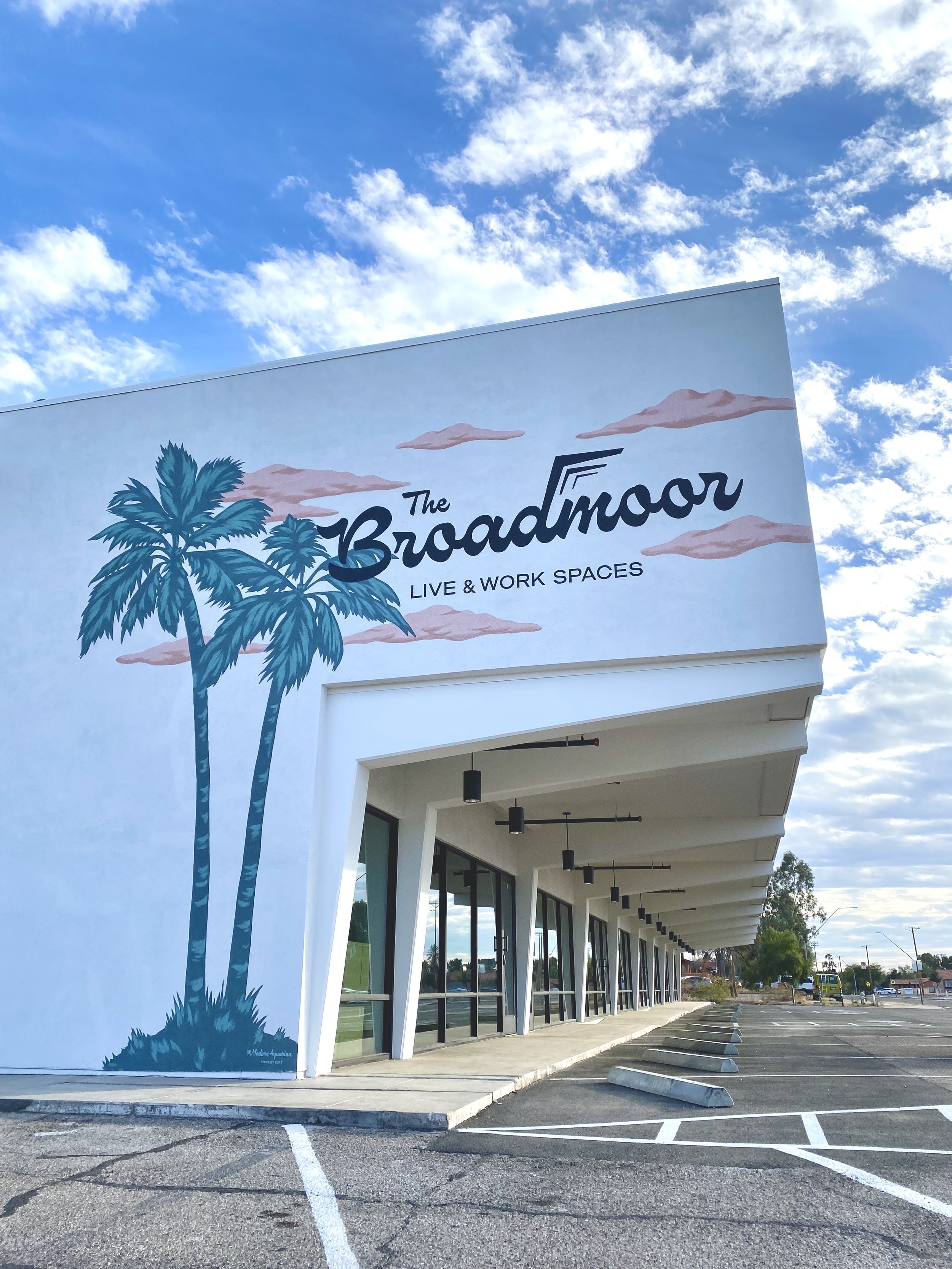 A commercial building with a large sign that reads 'The Broadmoor Live & Work Spaces' and features illustrations of palm trees and clouds on the exterior wall.