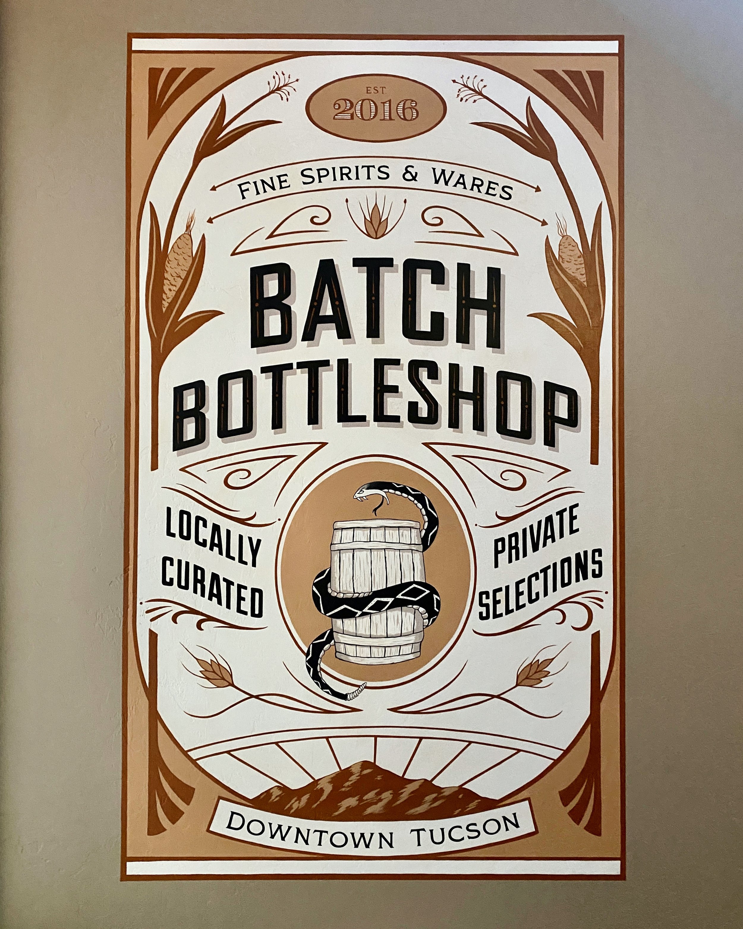 A poster advertising a batch bottling shop in downtown Tucson, established in 2016, offering locally curated private selections of fine spirits and wares, featuring a graphic of a snake coiled around a wooden barrel.