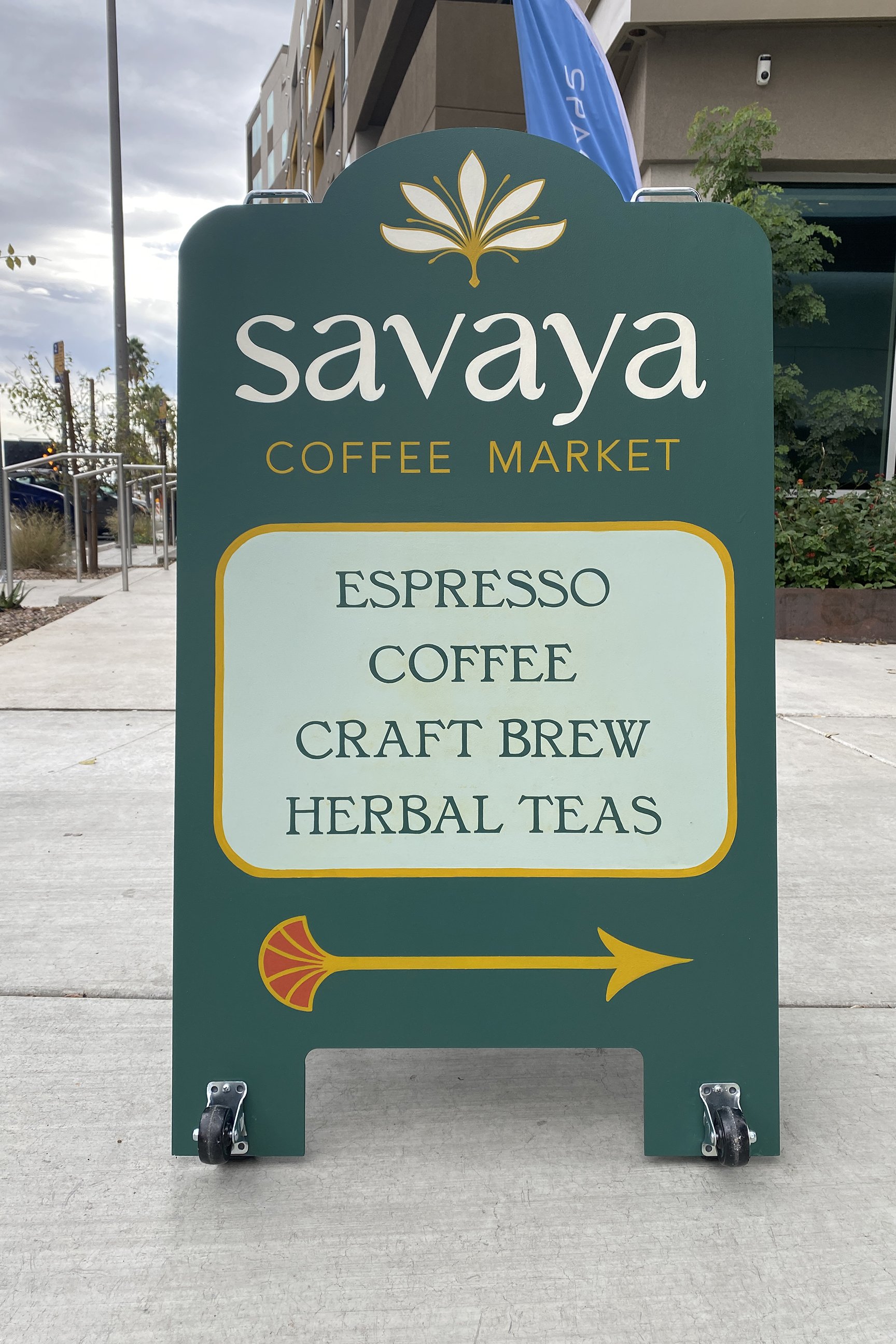 Green sidewalk signboard for Savaya Coffee Market listing espresso, coffee, craft brew, and herbal teas, with a yellow arrow pointing to the right.