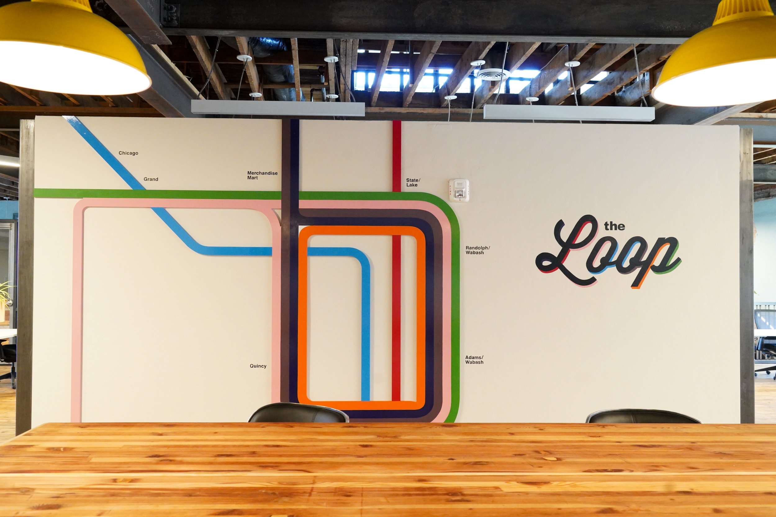 A map mural of a transit system on a wall behind a wooden table and two black chairs, with colored lines representing different routes and stations labeled, along with the text 'the Loop' written in colorful cursive.