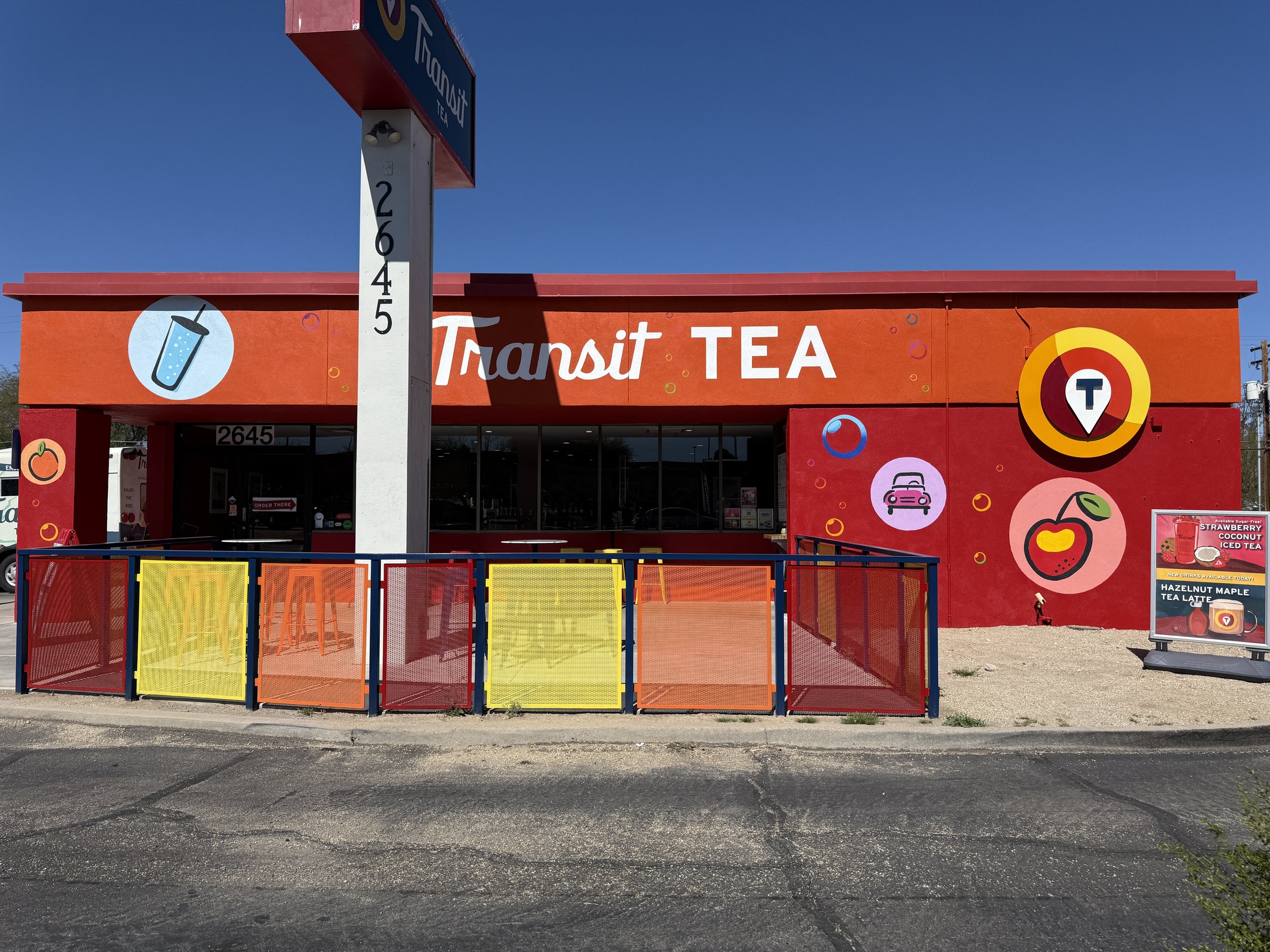Colorful storefront of Transit Tea with a bright red, orange, and yellow exterior, decorated with illustrations of a drink, cherry, peach, and car, and a parking lot in front.