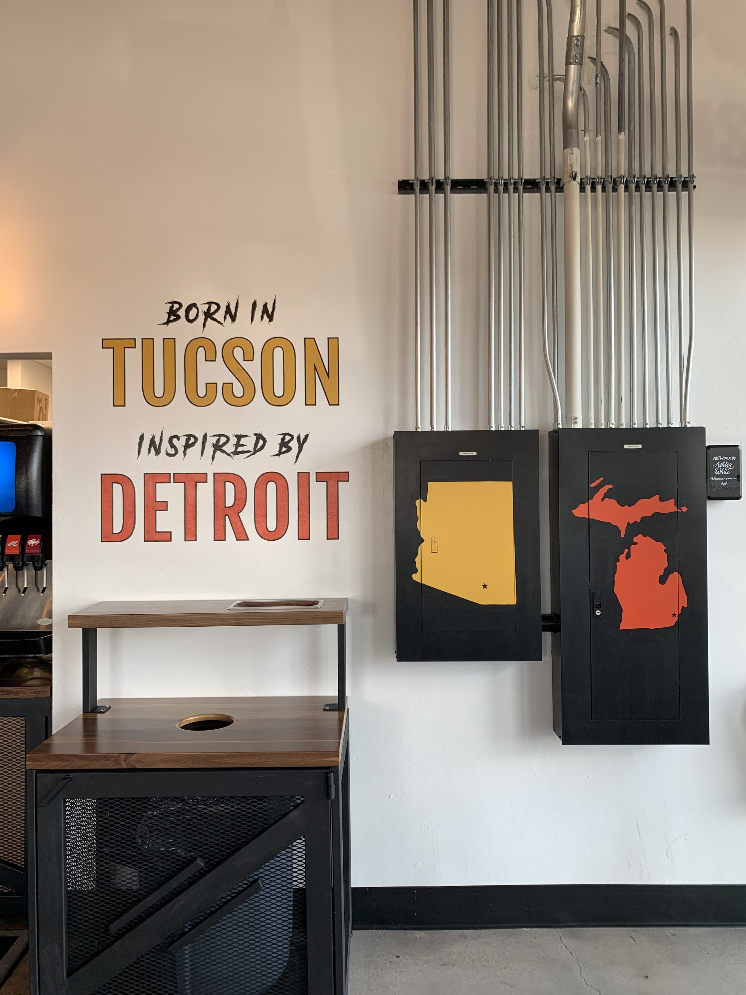 Wall with text 'Born in Tucson inspired by Detroit', two electrical cabinets with maps of Arizona and Michigan, and part of a beverage station on the left.