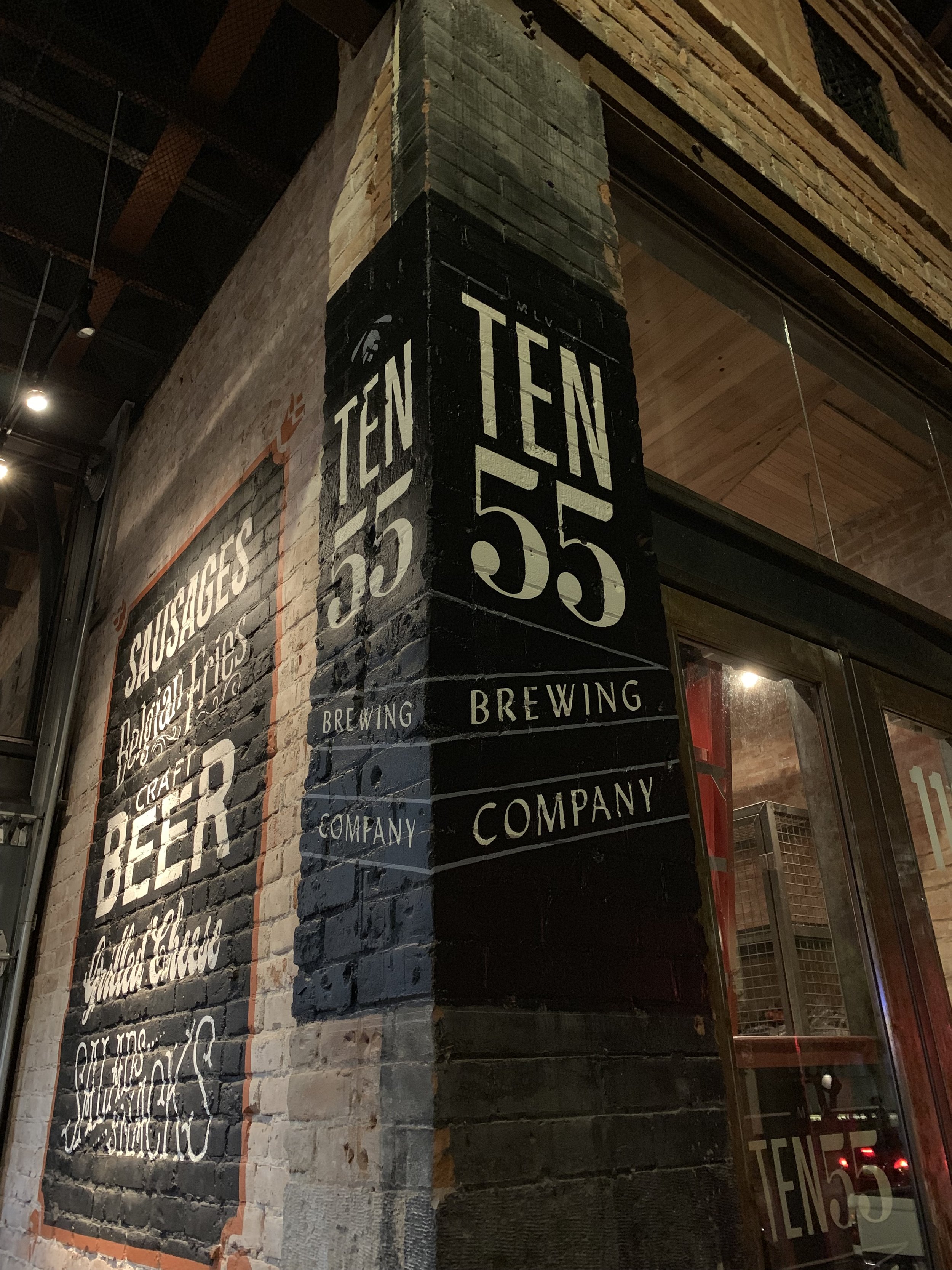 Brick wall with black and white painted signs for Ten55 Brewing Company and a menu for sausages and beer.