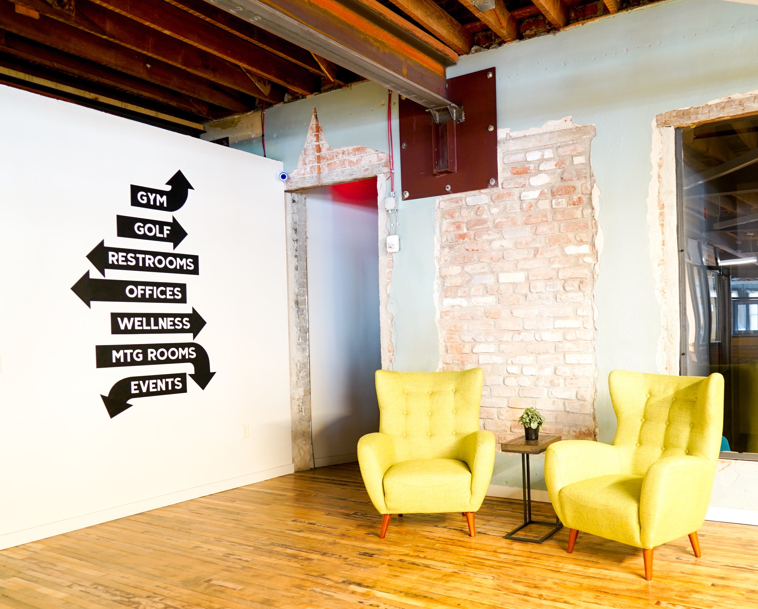 Interior view of a modern office space with two yellow armchairs, a small wooden table with a potted plant, a white wall with directional signs for gym, golf, restrooms, offices, wellness, MTG rooms, and events, exposed brickwork, and a ceiling with