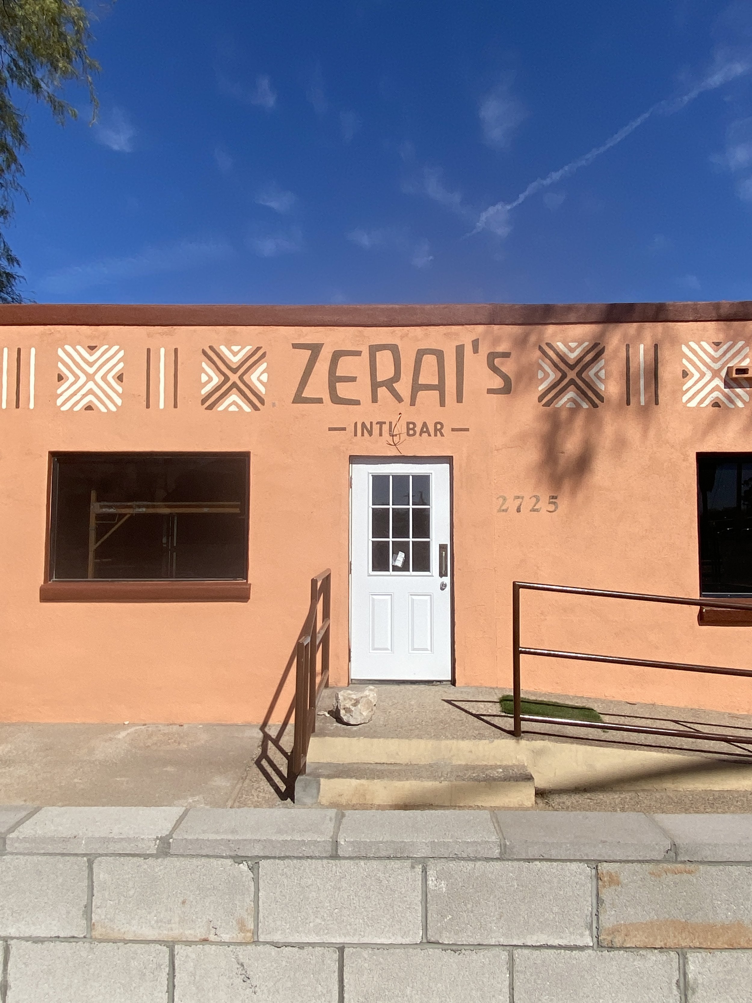 Exterior of a coral-colored building with a sign that reads 'Zerai's Inn & Bar,' featuring decorative patterns above the sign, a white door, a window to the left, and the number 2725 on the wall.