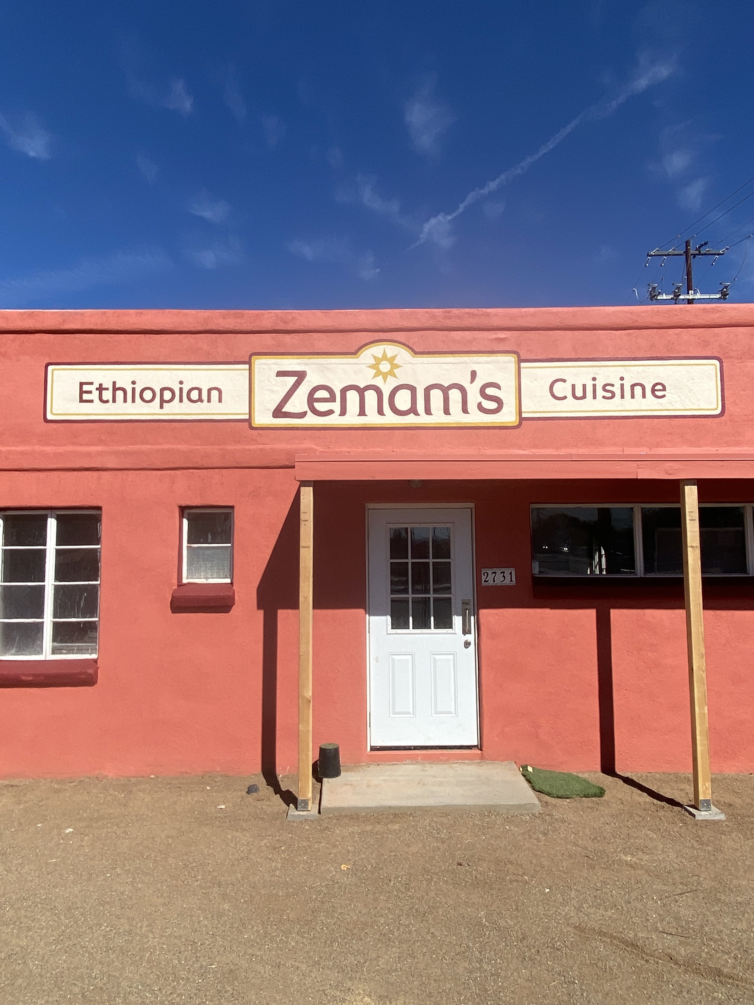 The storefront of Zemam's, a restaurant specializing in Ethiopian cuisine, painted in warm coral color with a white door, two windows, and a sign above reading 'Ethiopian Zemam's Cuisine.' The building is set against a clear blue sky.