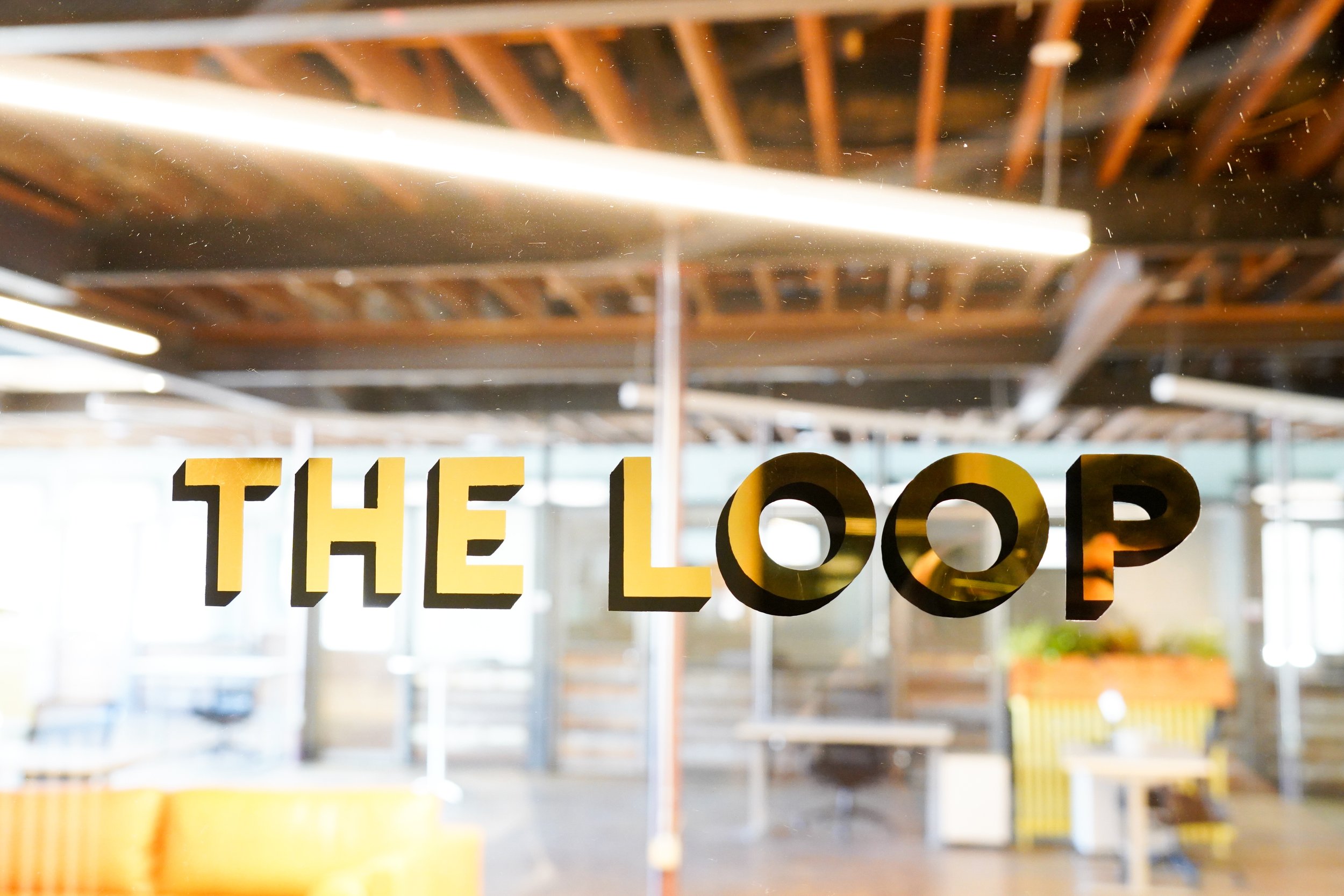 Sign that reads 'THE LOOP' on a glass window with a reflection of an industrial-style interior with exposed ceiling beams.