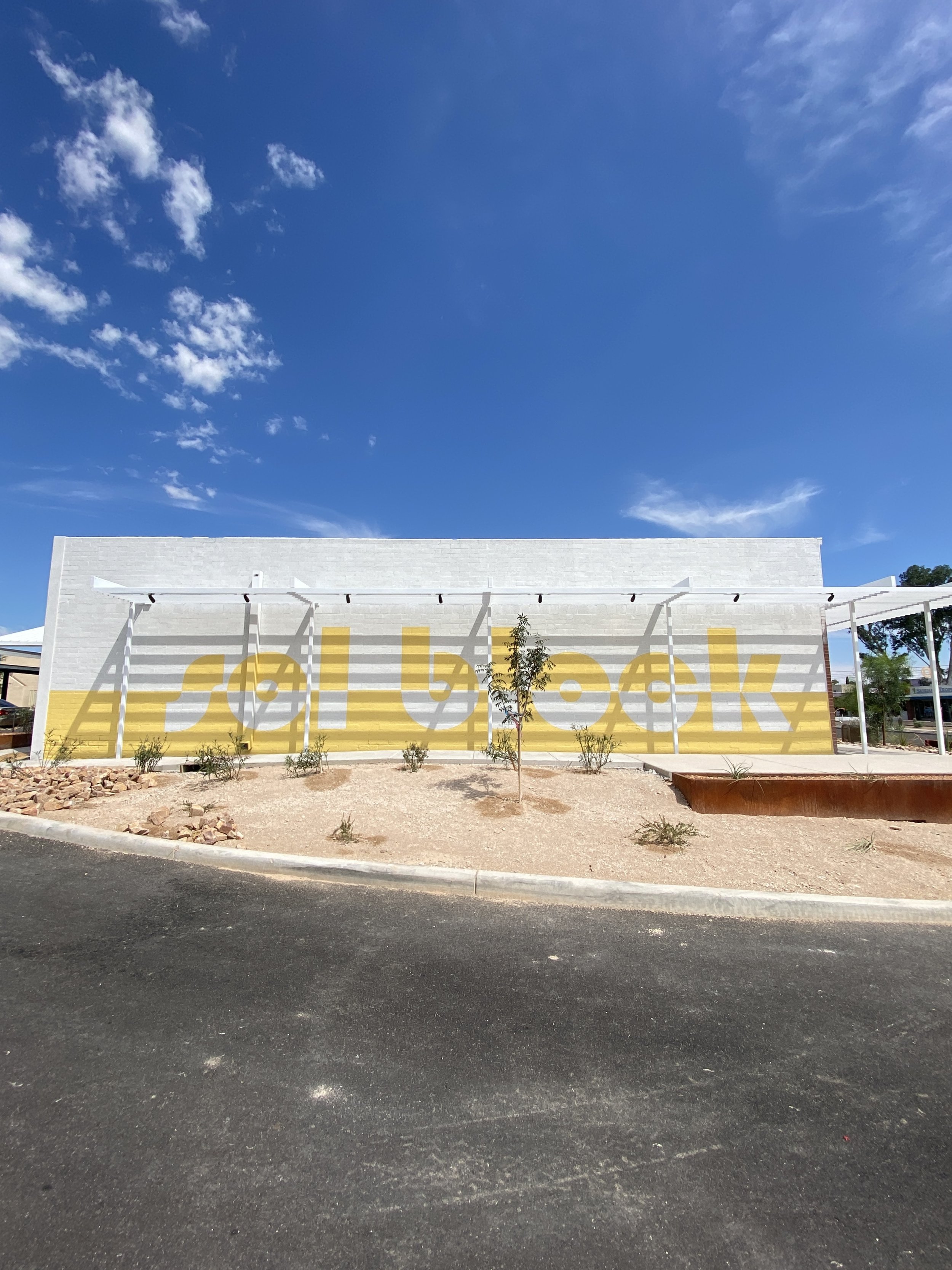 A white building with yellow graphics spelling out 'Solbook' on its facade, under a blue sky with scattered clouds, surrounded by small plants and a paved road in the foreground.