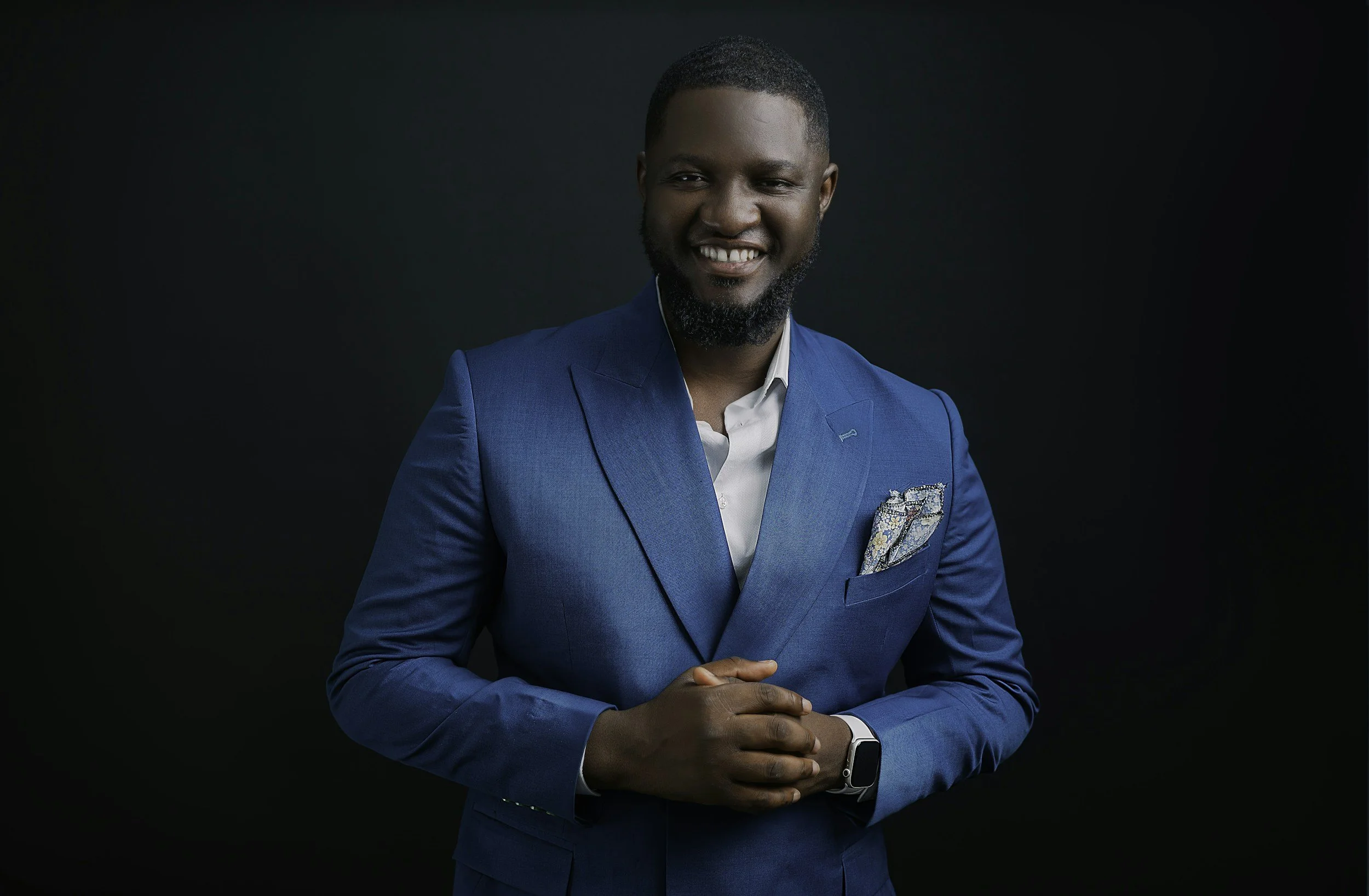 A smiling man in a blue suit with a pocket square, standing against a black background. He has a beard and short hair, and his hands are clasped in front of him.