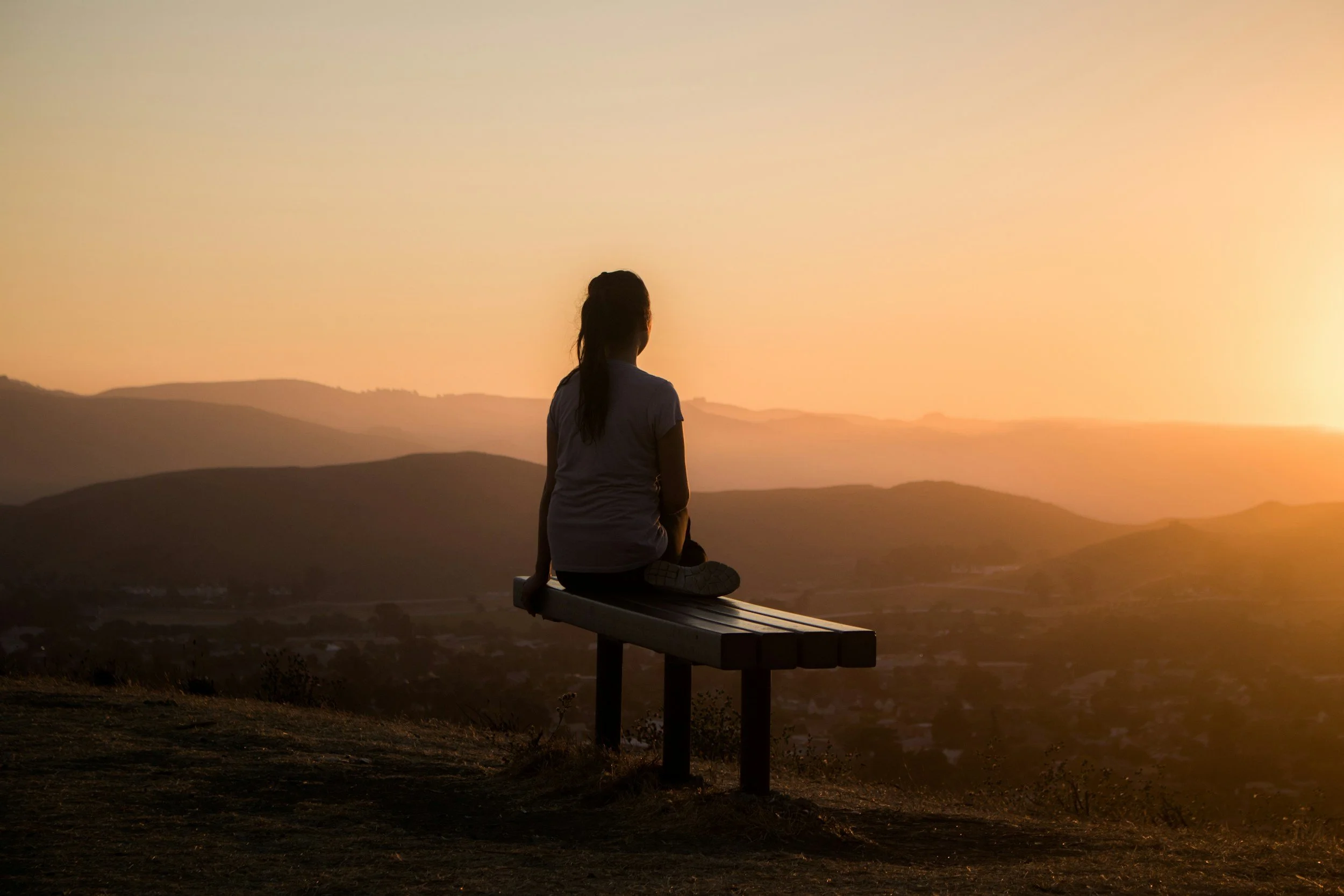 A woman with long dark hair sitting on a bench, looking at a sunset over distant hills and mountains.
