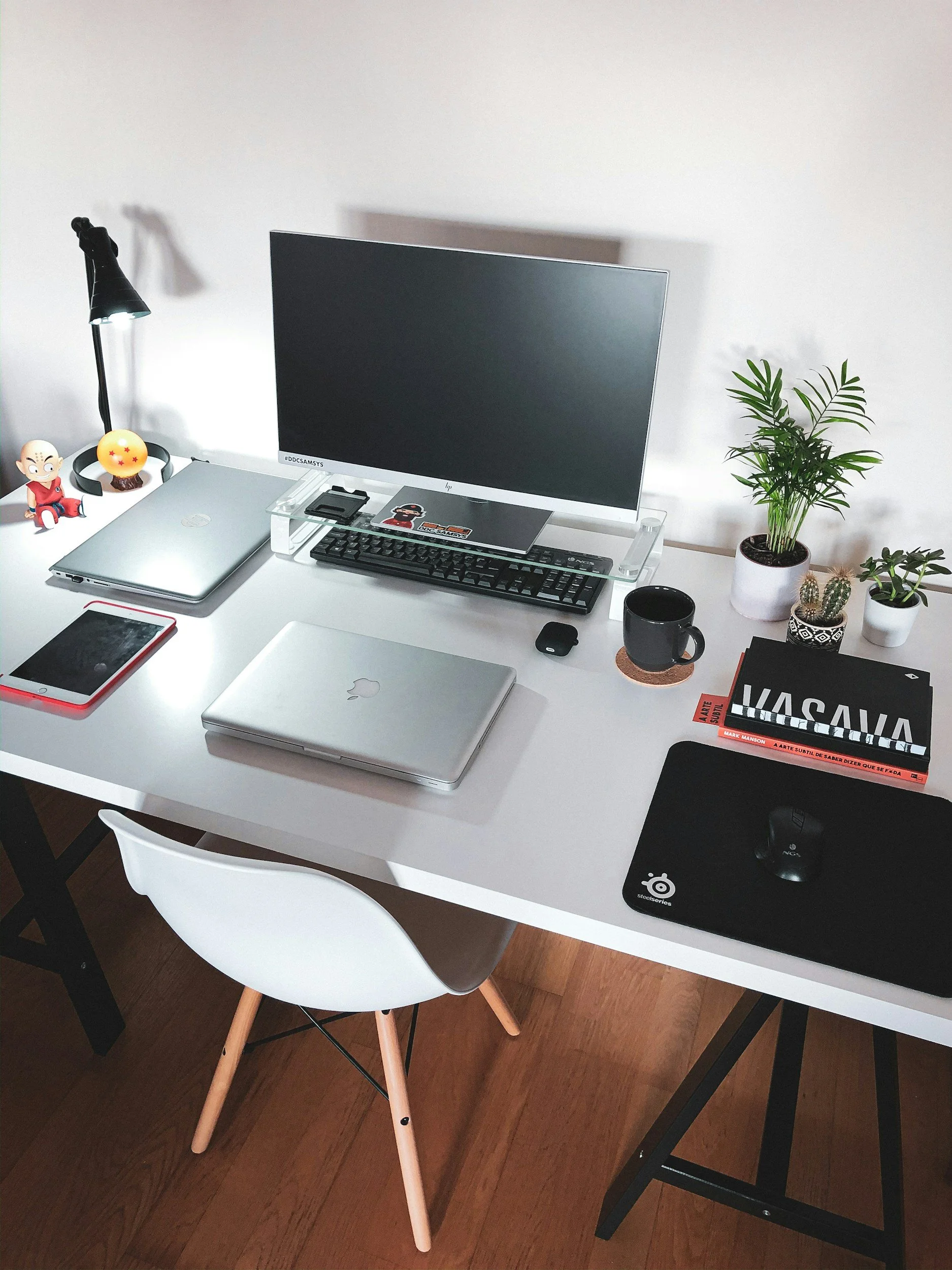 A white desk with a laptop, a tablet, a monitor, a black mug, a small black mousepad with a mouse, a keyboard, and several potted plants on the right side. There is a white chair with wooden legs in front of the desk. The background shows a white wall.