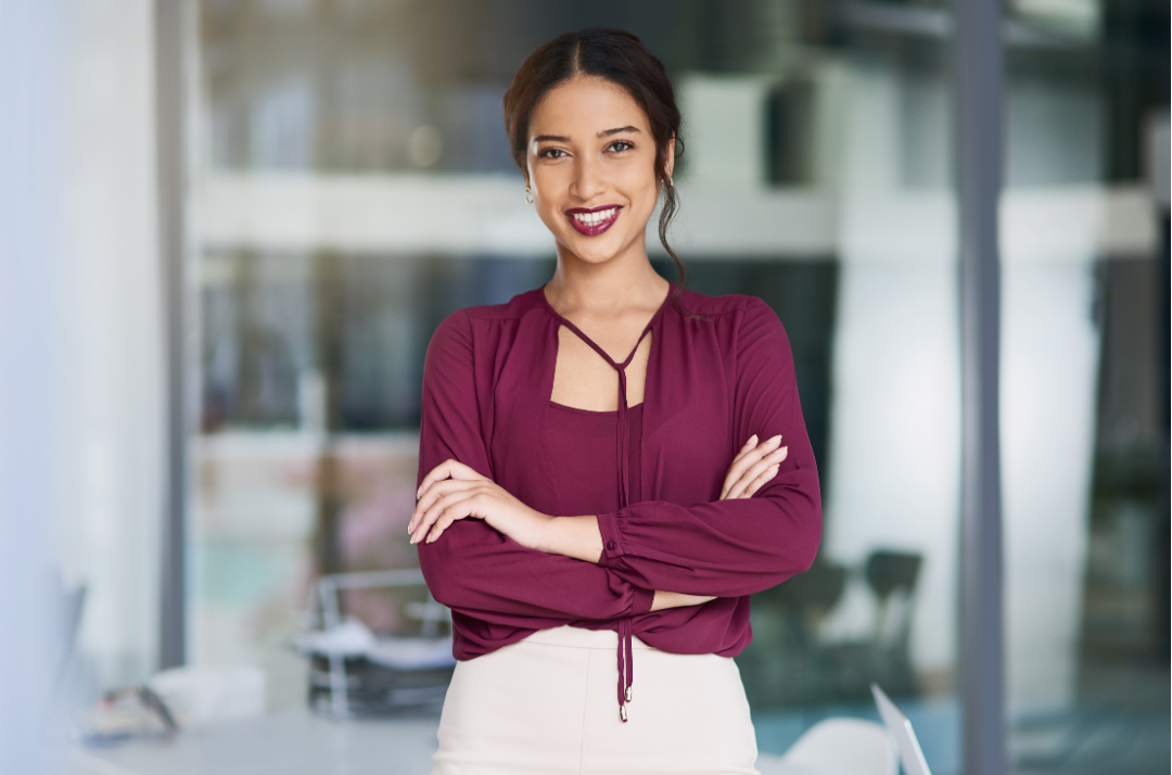 A woman wearing a burgundy blouse and cream skirt smiling with arms crossed in an office setting.