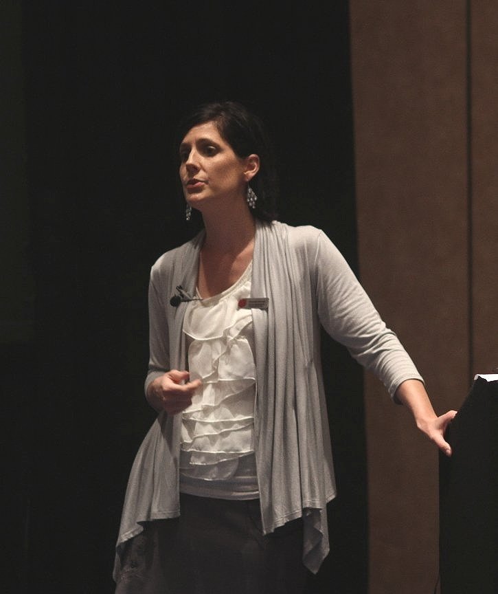 A woman with dark hair, wearing earrings and a light-colored cardigan, standing in a room with a dark and wood-paneled background, possibly giving a presentation or speech.