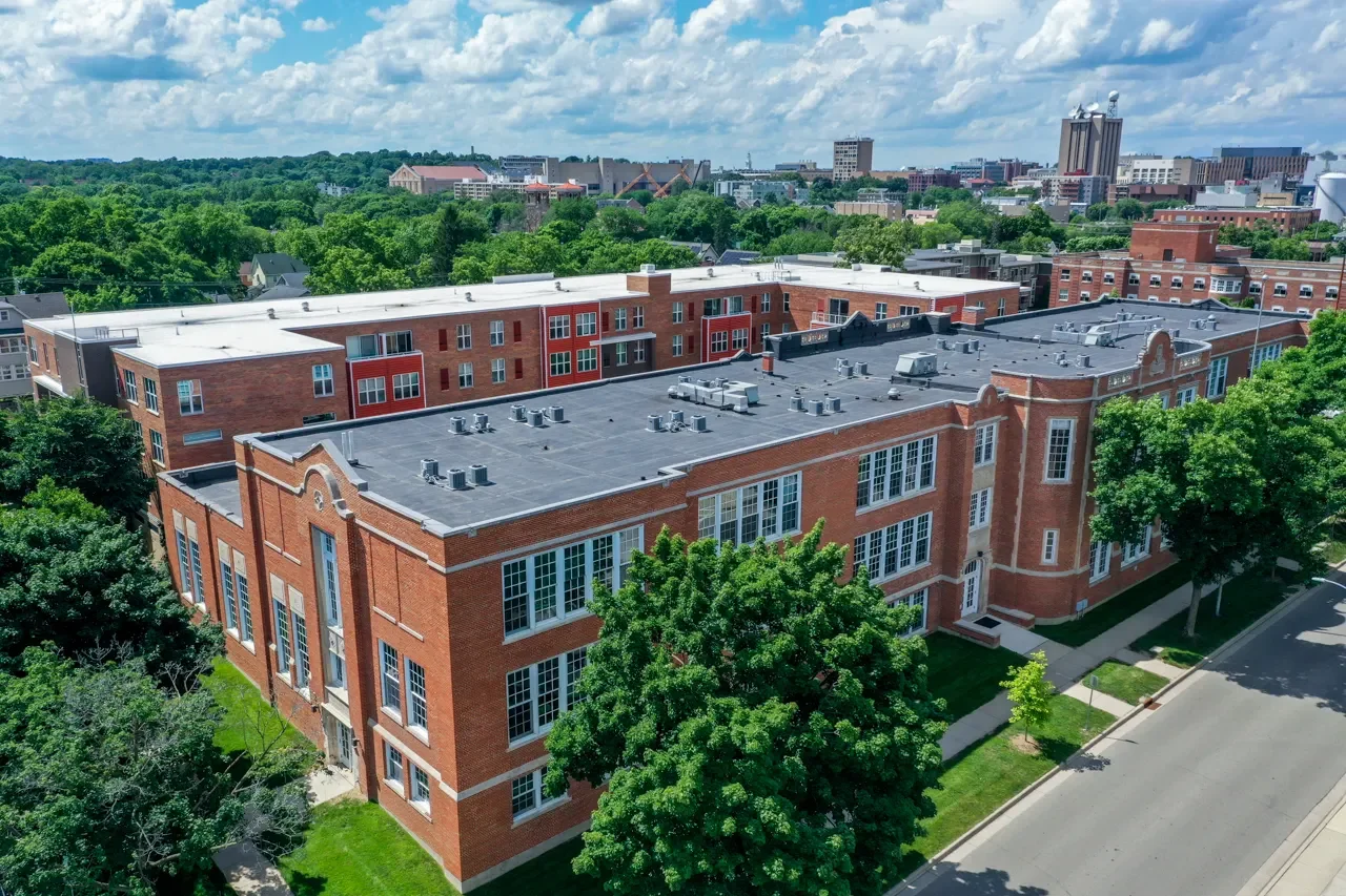 Aerial view of a red brick building with large windows and surrounding trees in a city.