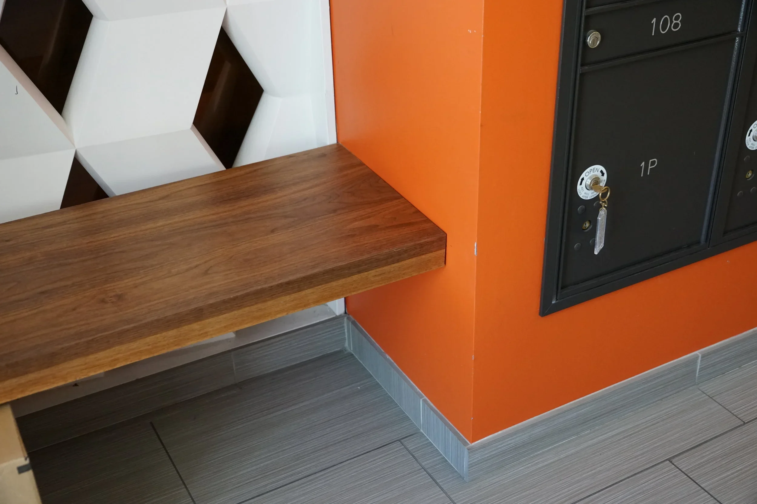 Corner of a room with a wooden countertop, a black mailbox with a keyhole, and a bright orange wall. Two white chairs can be seen in the background.