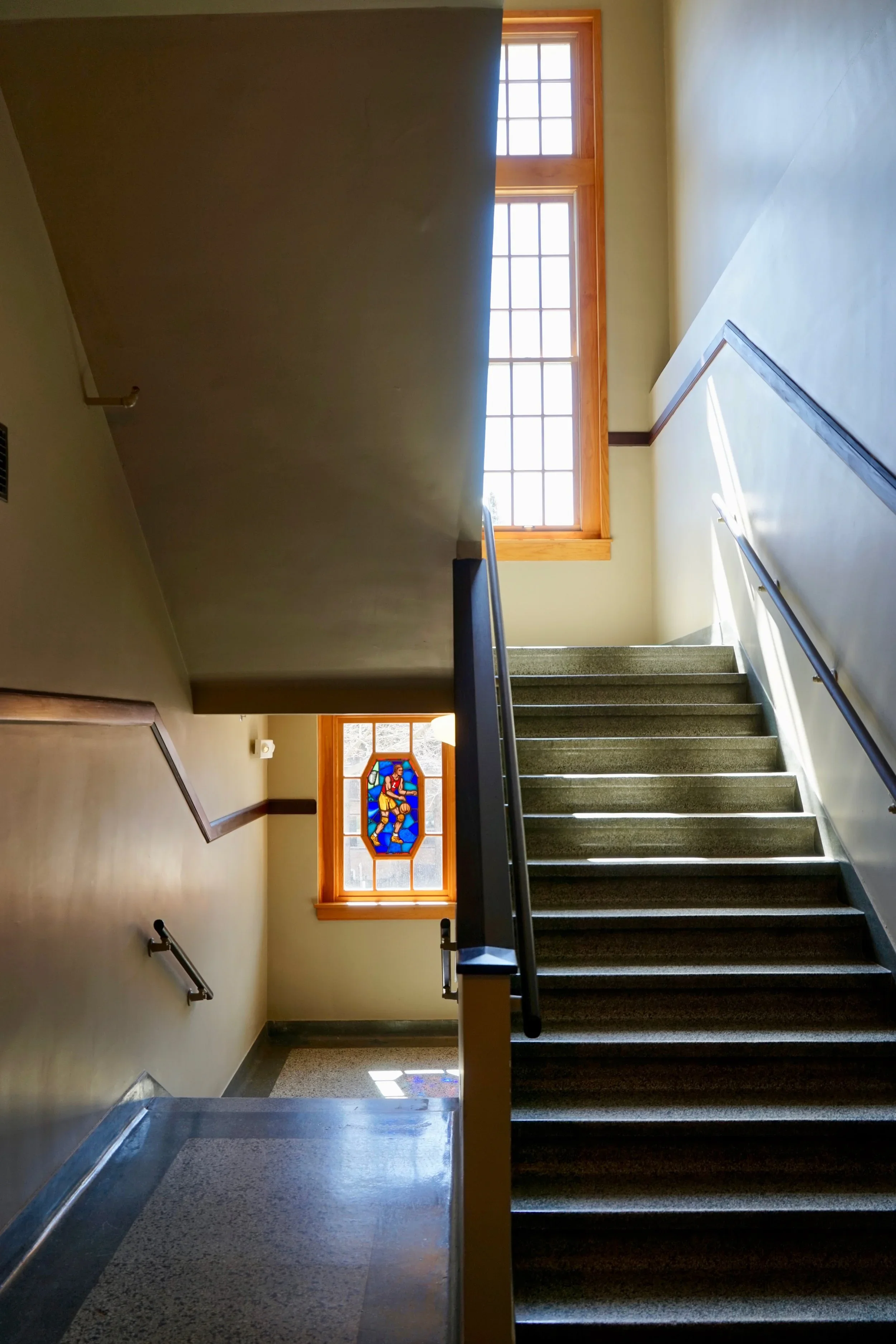 Indoor staircase with green carpet and handrails, leading up to large windows with wooden frames and a stained glass window below the larger window, allowing sunlight to illuminate the hallway.