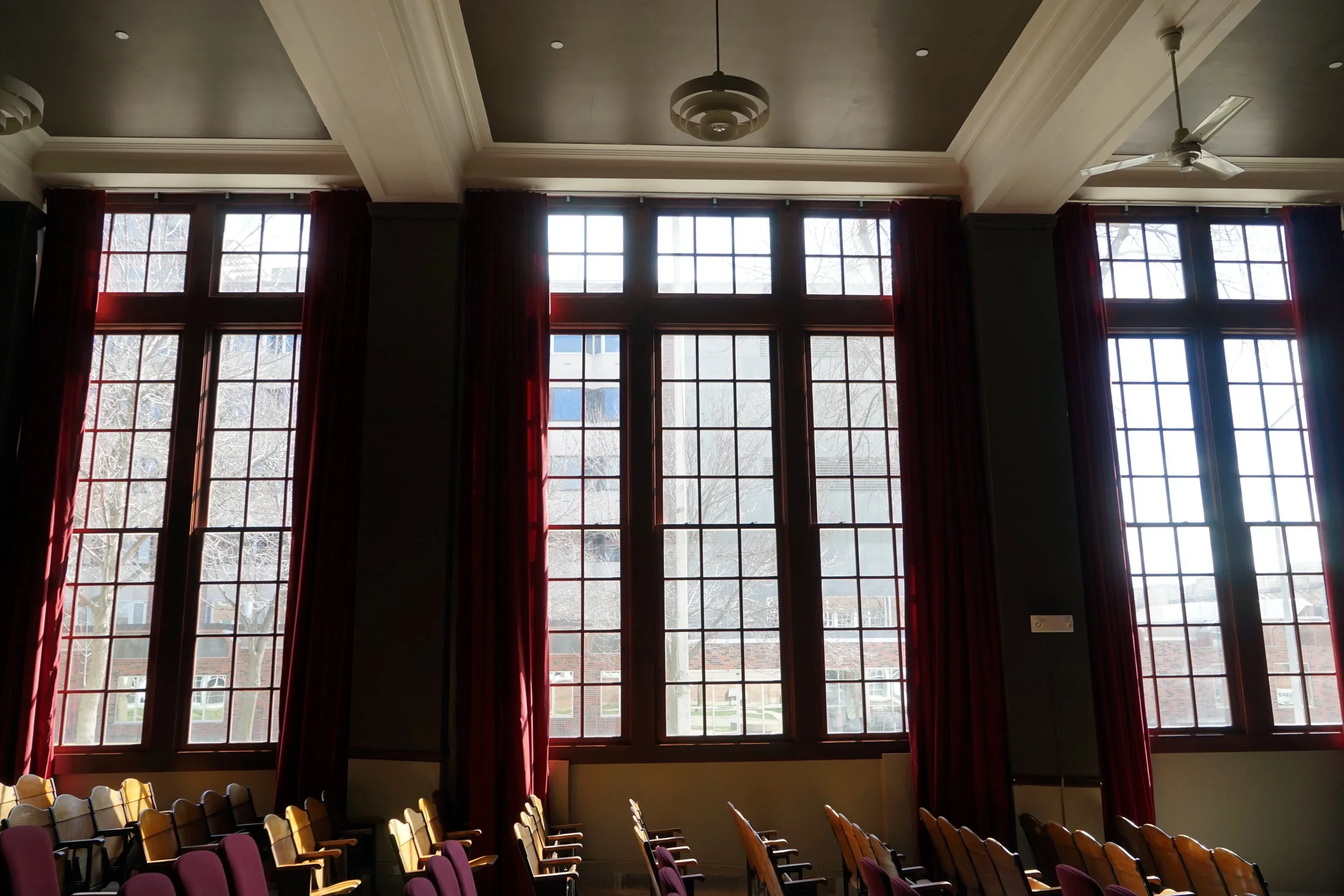 Interior of a room with large windows, red curtains, ceiling fan, and a chandelier. Rows of chairs are arranged along the walls, facing a stage or presentation area.