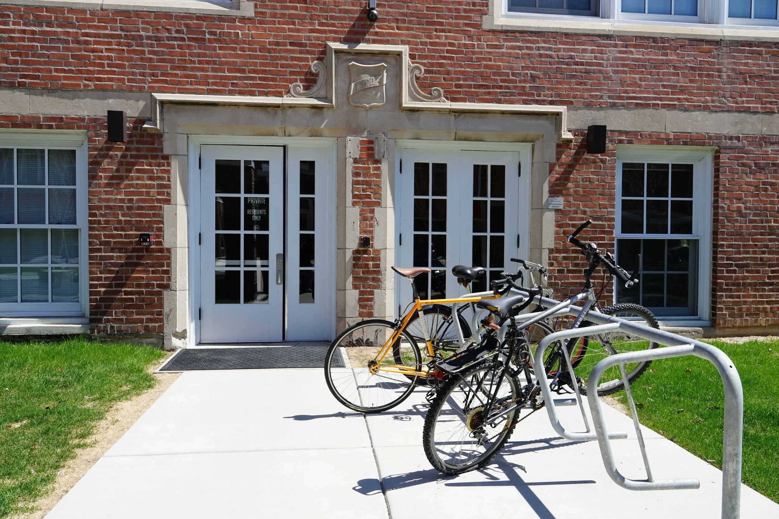 Brick building with two white doors, bicycle racks with bikes, and bikes parked in front, green grass on the side, and a window above.