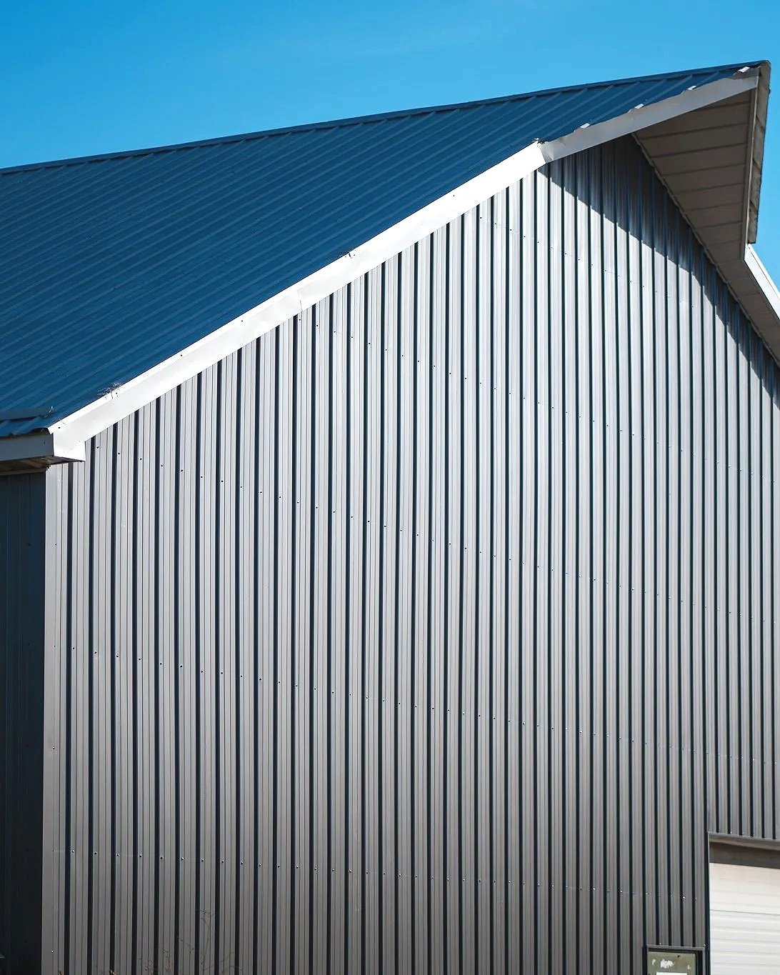 Close-up of a modern building with silver corrugated metal siding and a blue metal roof, under a clear blue sky.