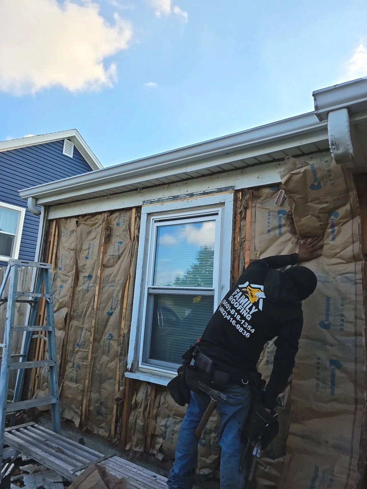 A construction worker in black clothing and a black beanie installing or repairing the exterior wall of a house, with insulation and window frame visible. Ladder present nearby. Blue siding house can be seen in the background under a partly cloudy sky.