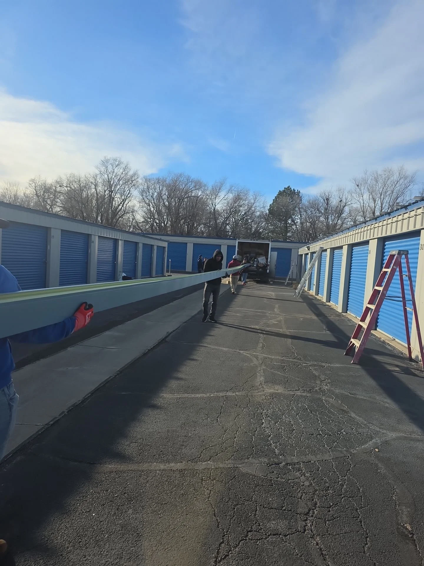 People working in a storage facility with blue roll-up doors, assembling a long piece of material, possibly a boat or similar object, in an outdoor lot with a partly cloudy sky and leafless trees in the background.