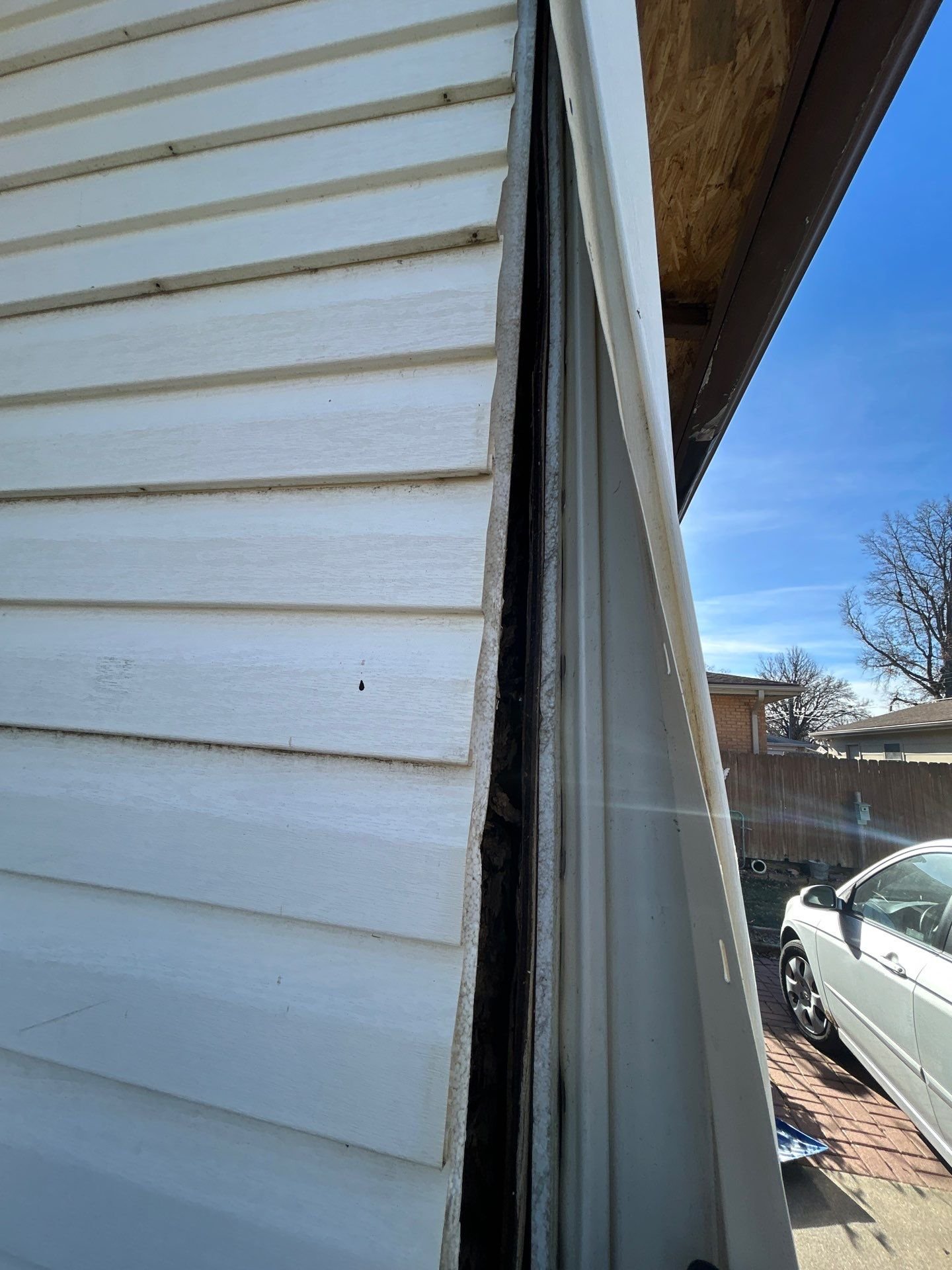 Close-up of damaged white siding and window frame on a house, with a view of a driveway, parked car, and clear blue sky.