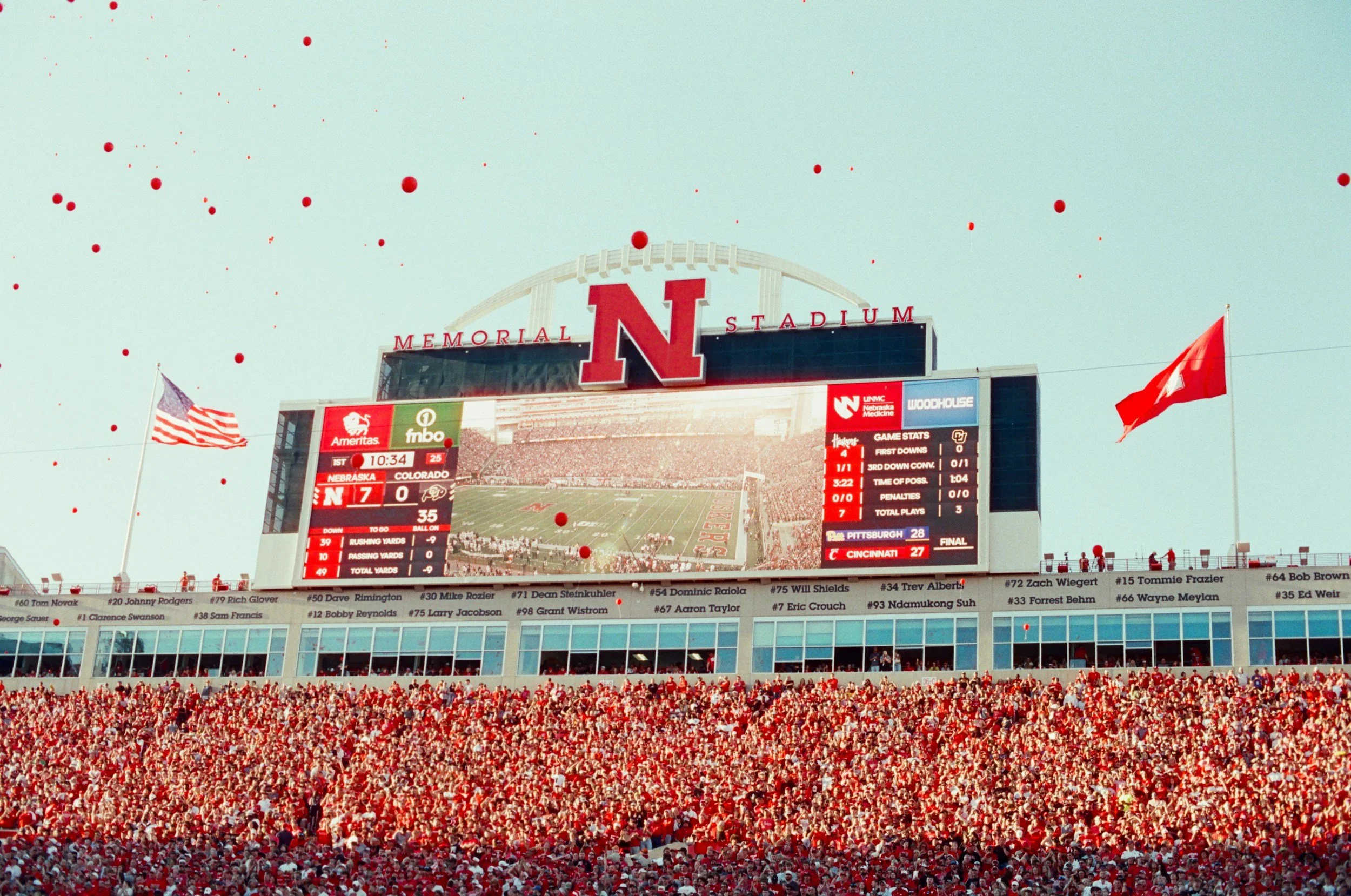 View of Memorial Stadium filled with fans, displaying a large scoreboard showing a football game between Nebraska and Colorado, with red balloons in the sky and flags waving.