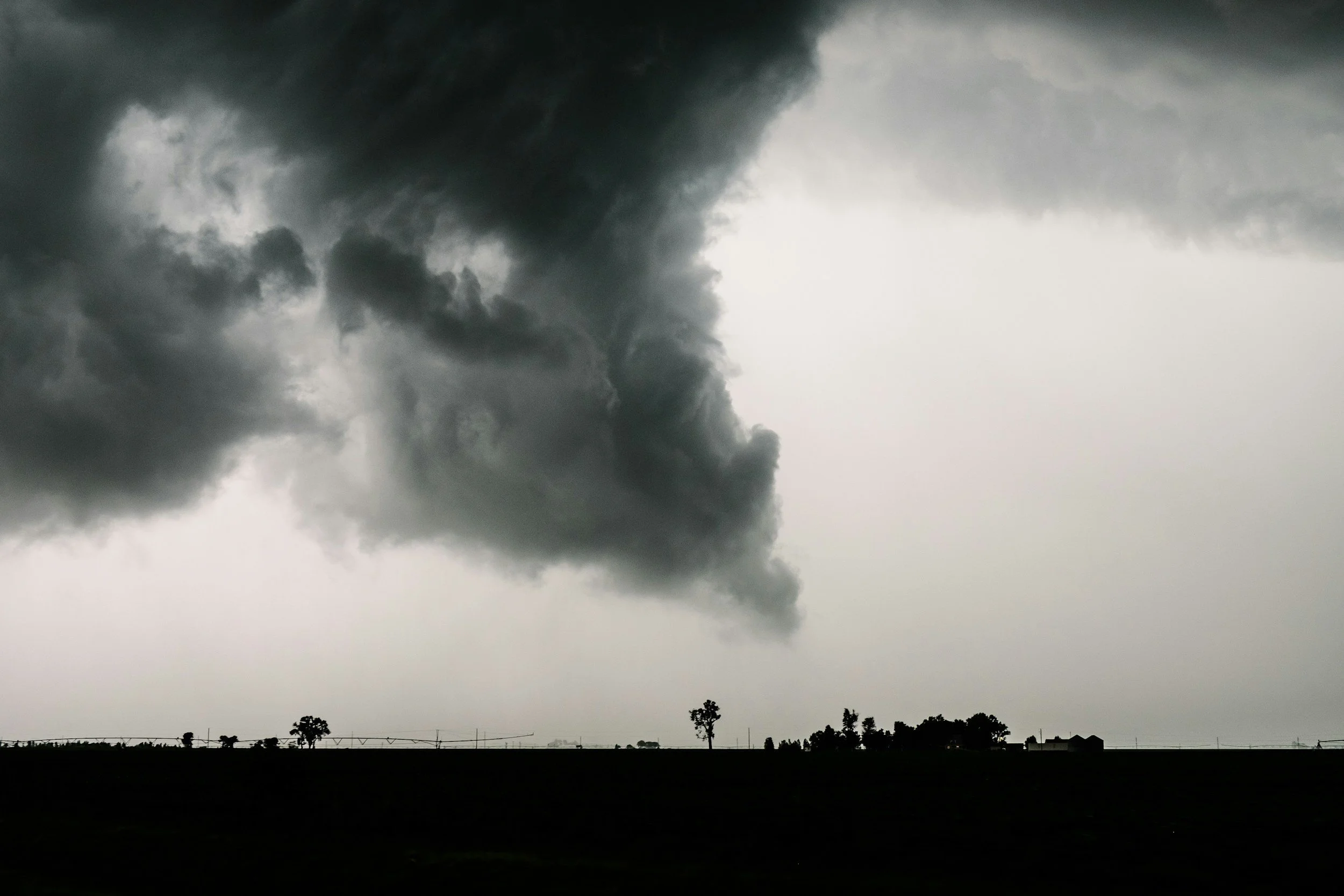 Dark storm clouds over a rural landscape with a few trees and houses in the distance.