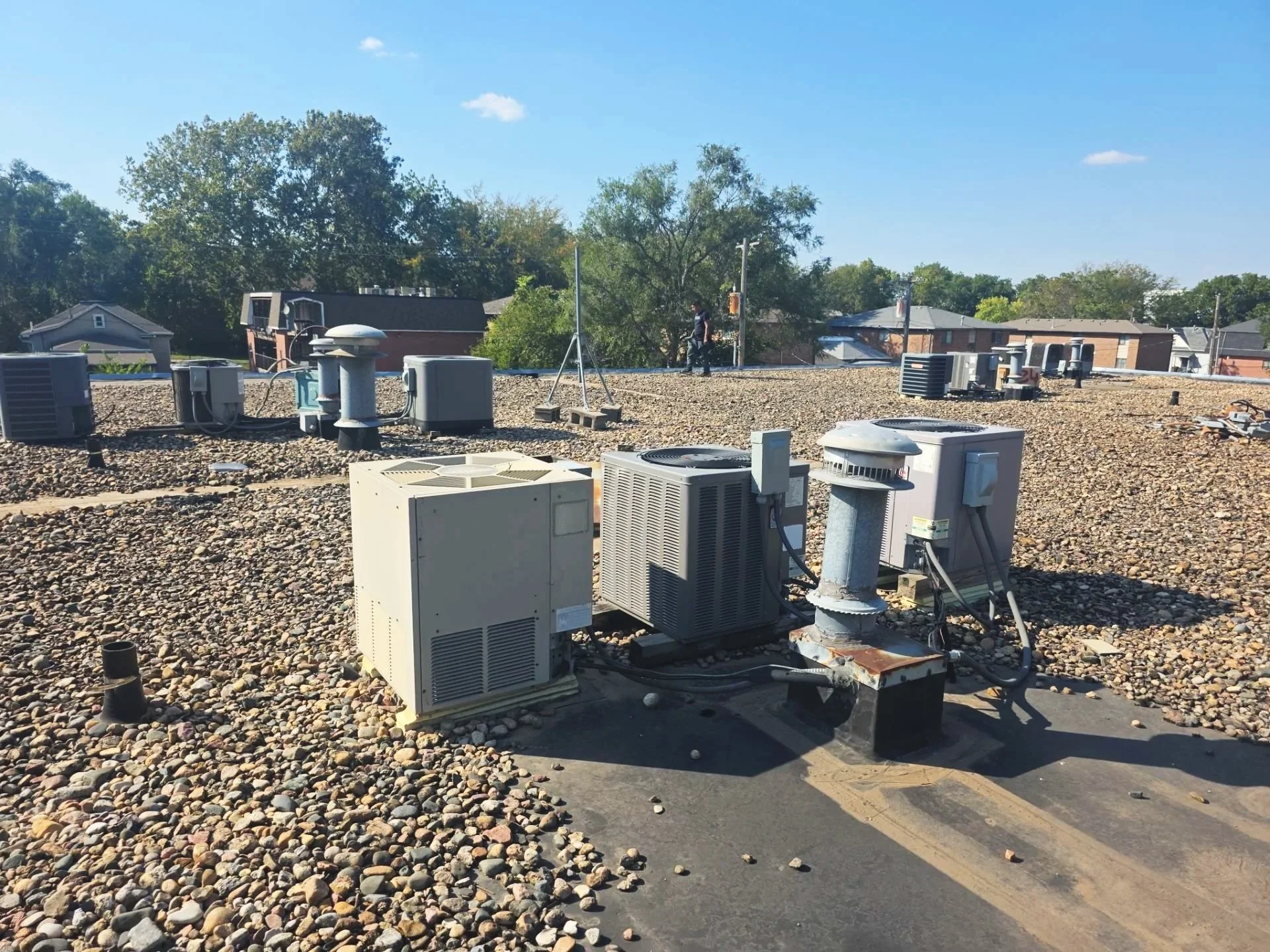 A rooftop with multiple HVAC units and vents on a clear, sunny day. Residential houses and trees are visible in the background.