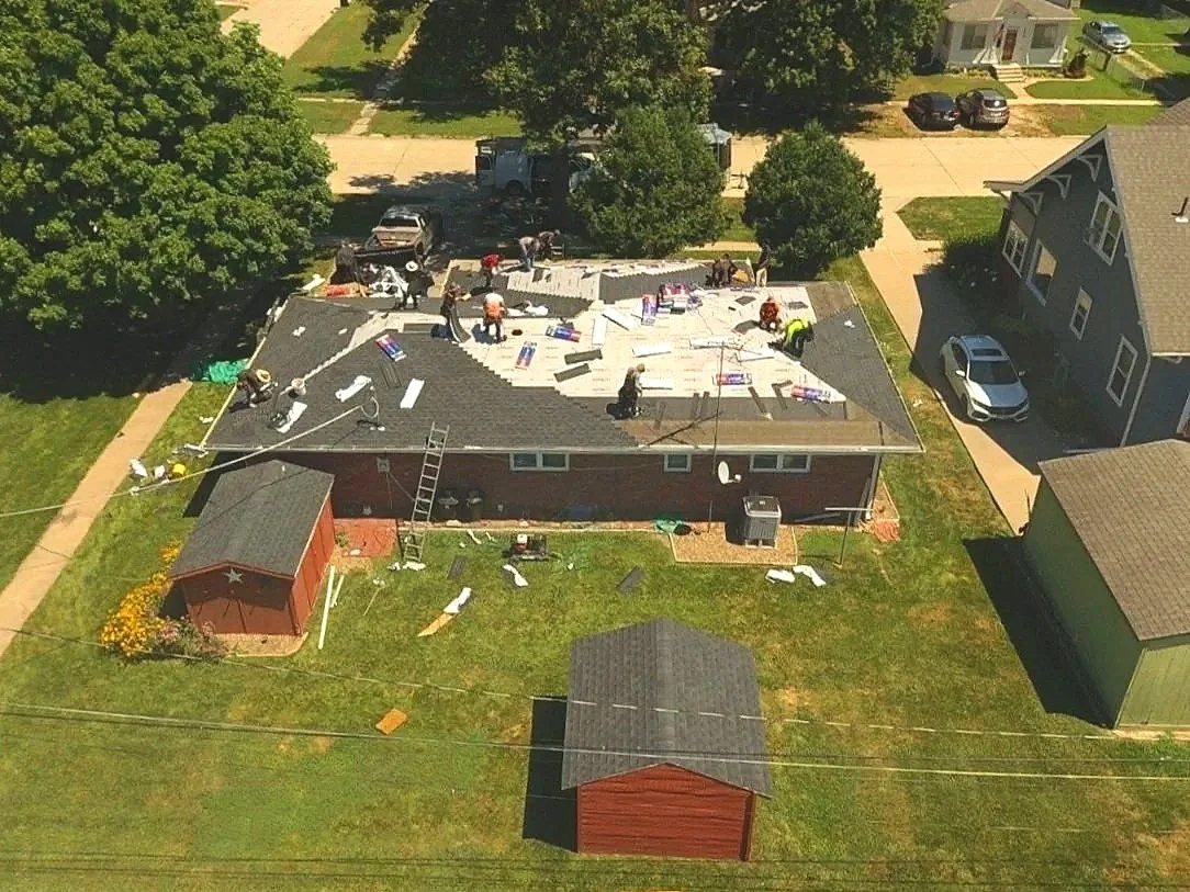 Aerial view of a house with a large section of the roof being repaired or replaced, with workers present. Tools and materials are scattered around, and there is a small red shed and a detached garage in the yard.