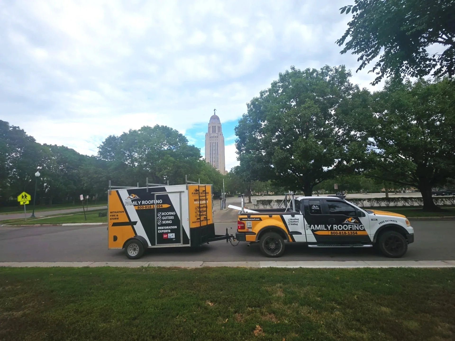 A truck with a trailer parked on a street with trees in the background and the Nebraska State Capitol building in Lincoln, Nebraska visible in the distance.