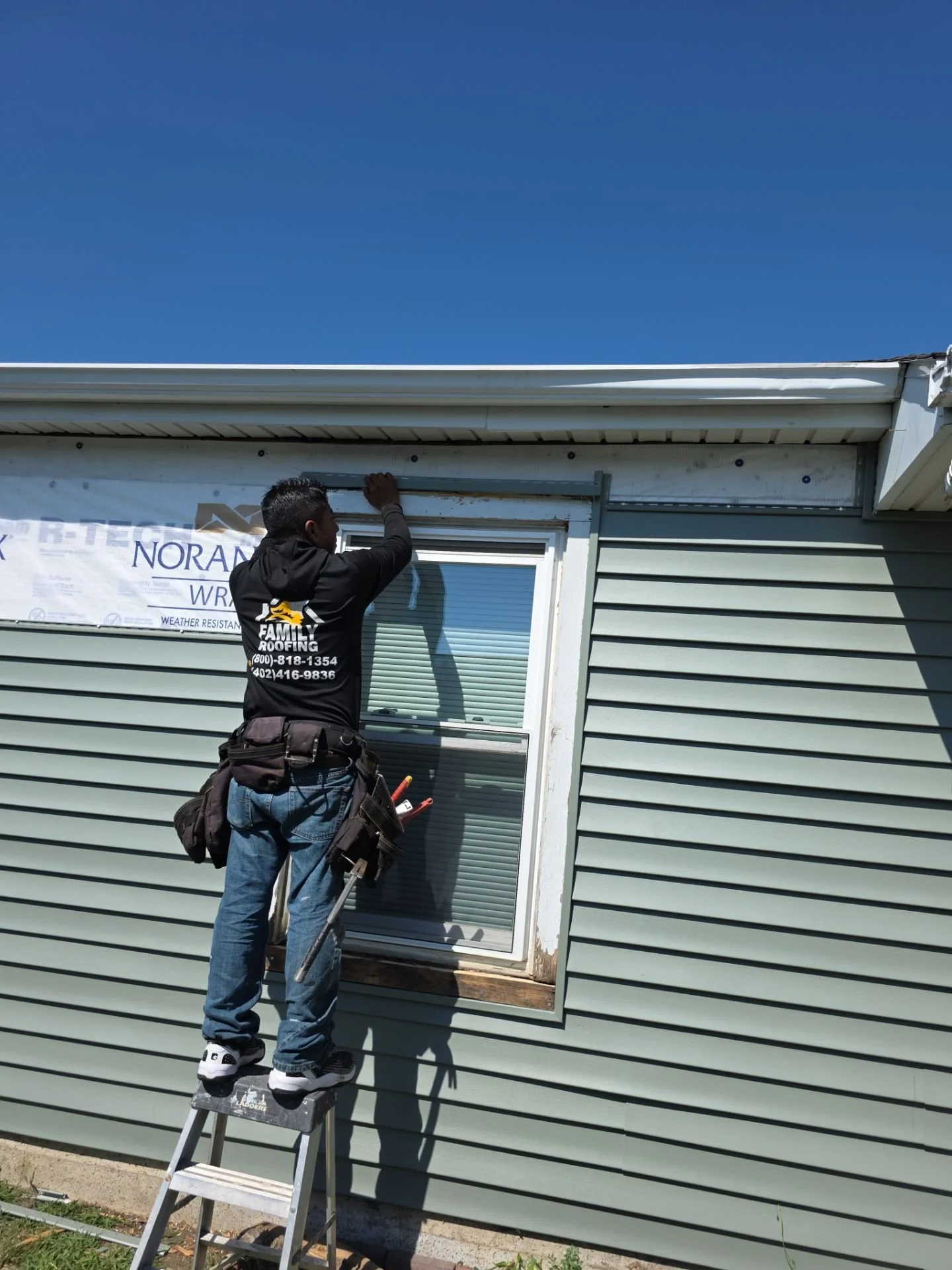 A man standing on a ladder installing or repairing a window on the exterior of a house with green siding, under a clear blue sky.