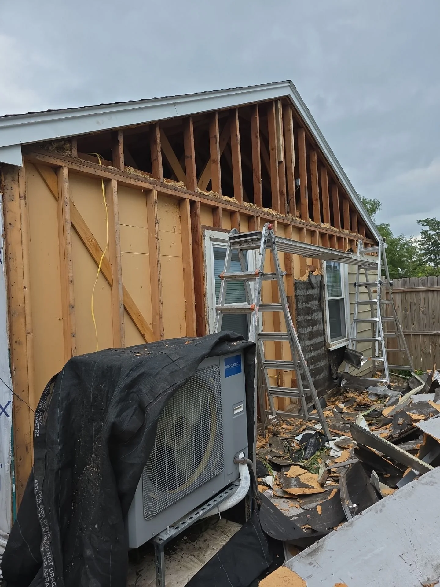 House under construction with exposed wooden framing and part of the exterior wall removed. Two ladders leaning against the house, debris on the ground, and an air conditioning unit covered with a black tarp.