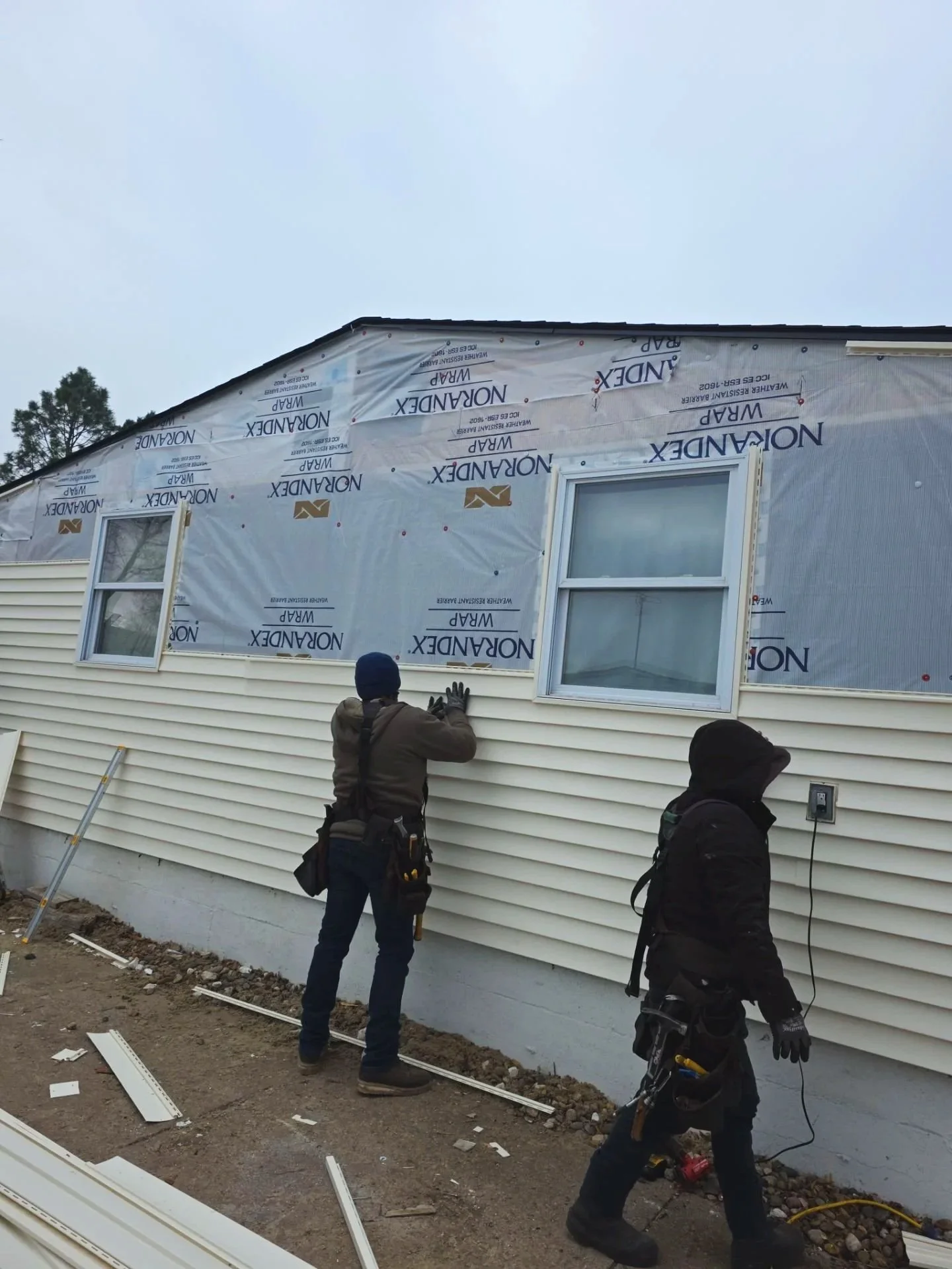Two construction workers installing siding on a house, with one worker near the wall and the other holding a power cord.